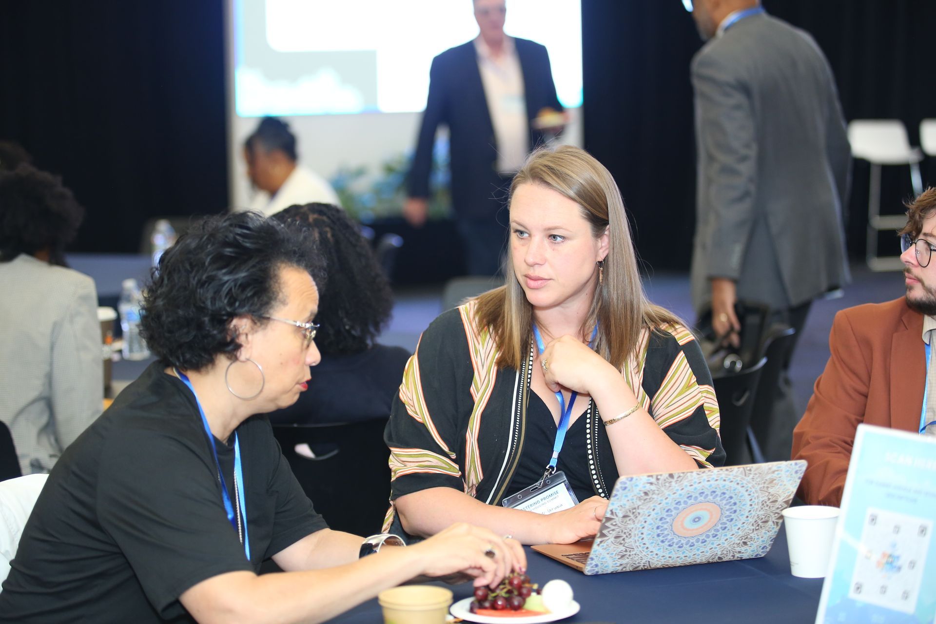 Two people talking at a table during a conference, with a laptop and coffee cup nearby.