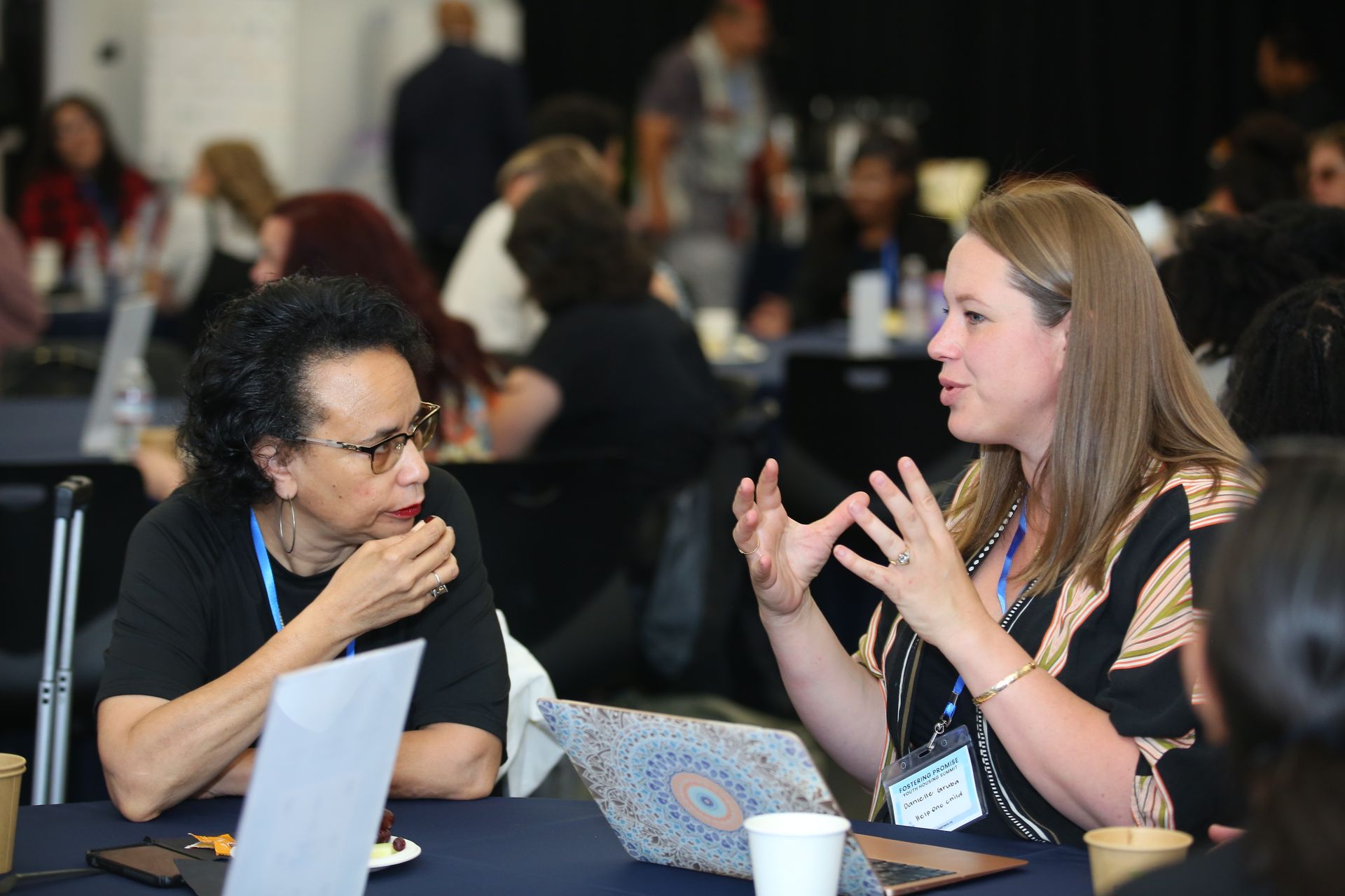 Two people talking at a table with laptops in a busy conference setting
