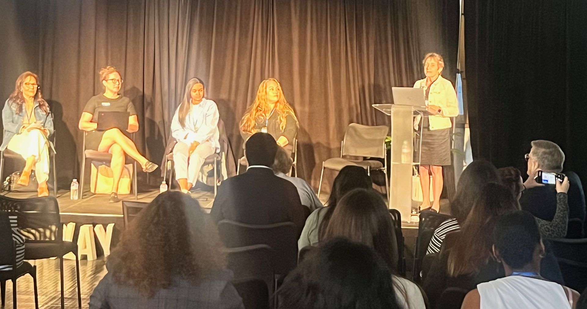 Panel discussion on stage with five speakers and audience seated in a dimly lit room