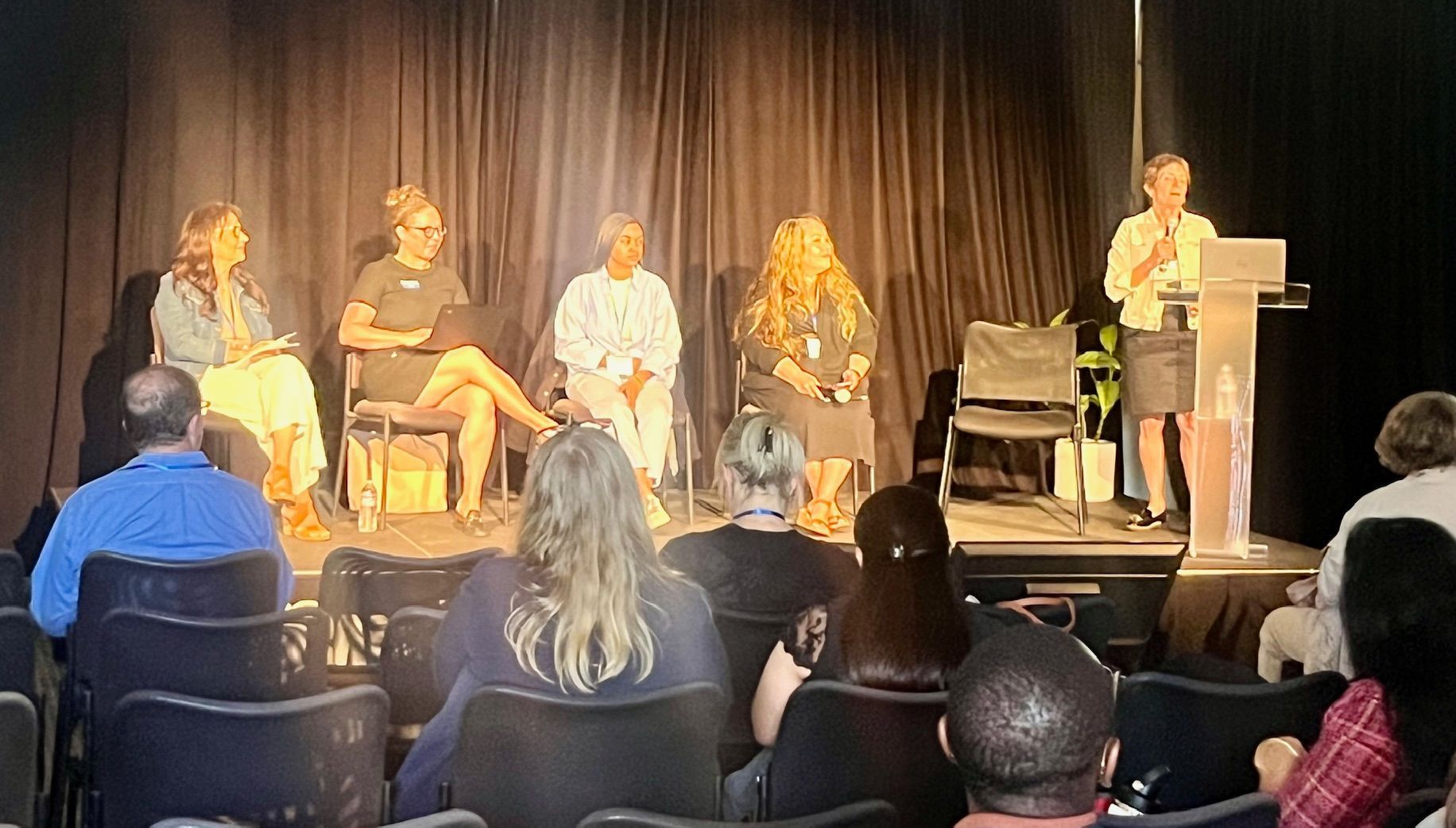 Panel discussion on stage with five seated speakers and a presenter at a podium in front of an audience