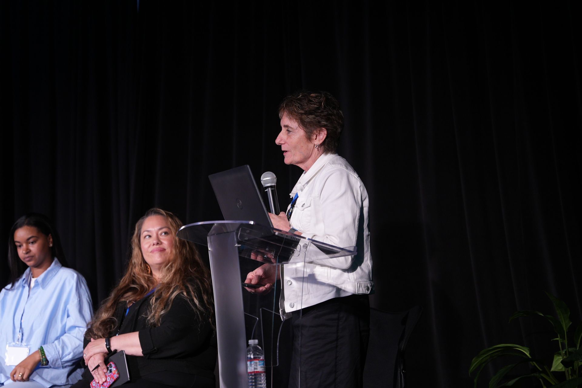 Speaker at podium on a dark stage, with two seated panelists beside her and a microphone nearby