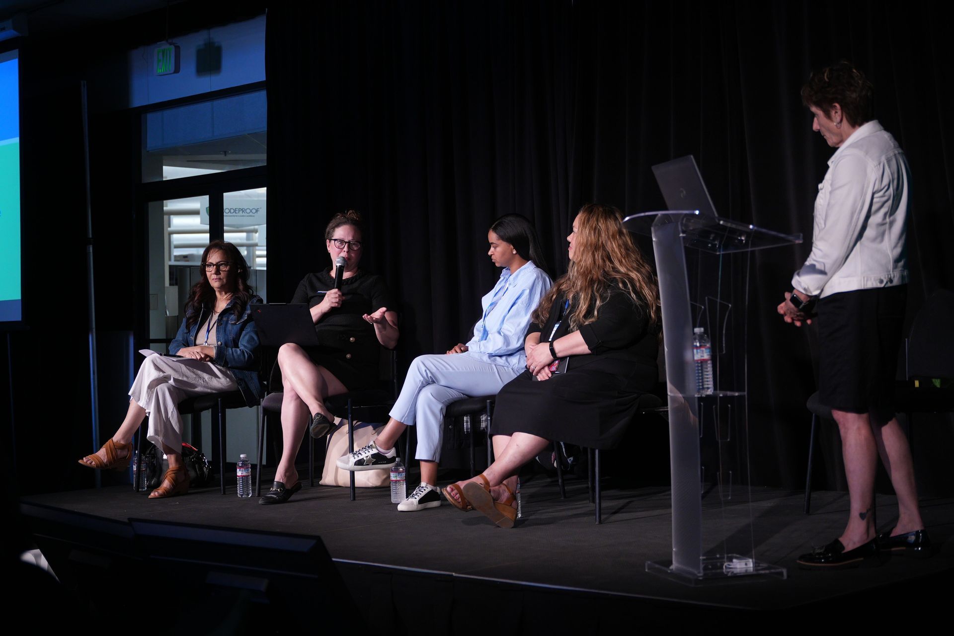Panel discussion on a dark stage, with four seated speakers and a moderator at a clear podium.