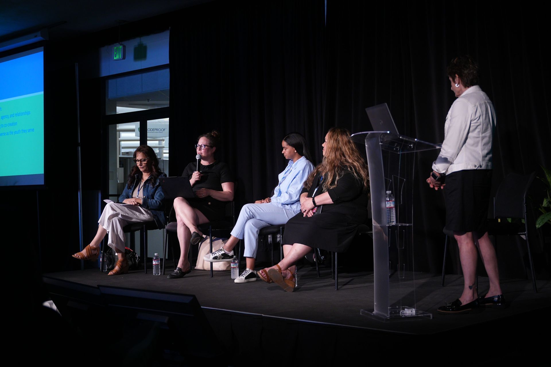 Panel discussion on a stage, with five seated speakers and a presenter at a podium, projected slides in the background