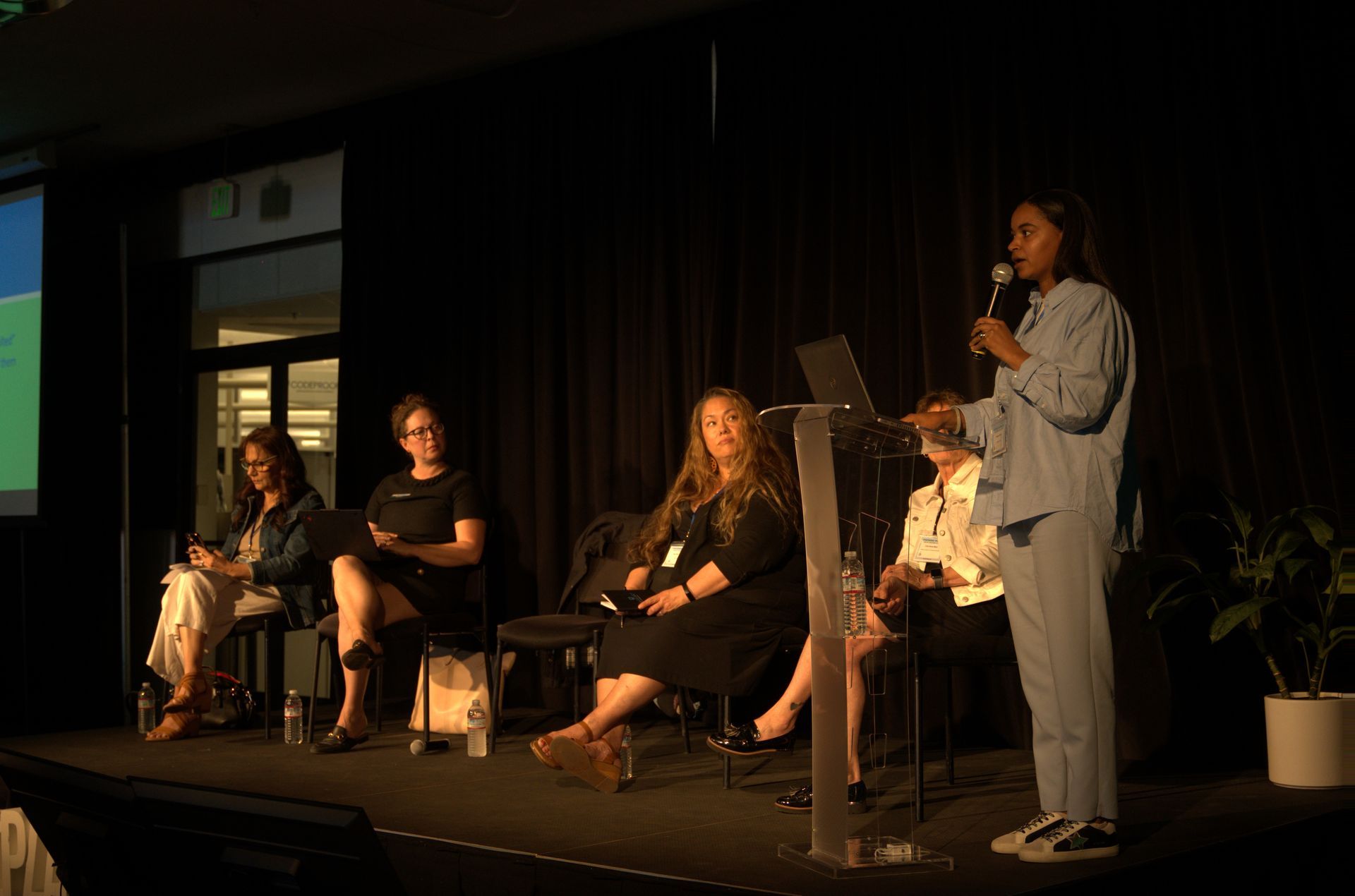 Panel discussion on a dark stage, with one speaker standing and four seated panelists under warm lights.