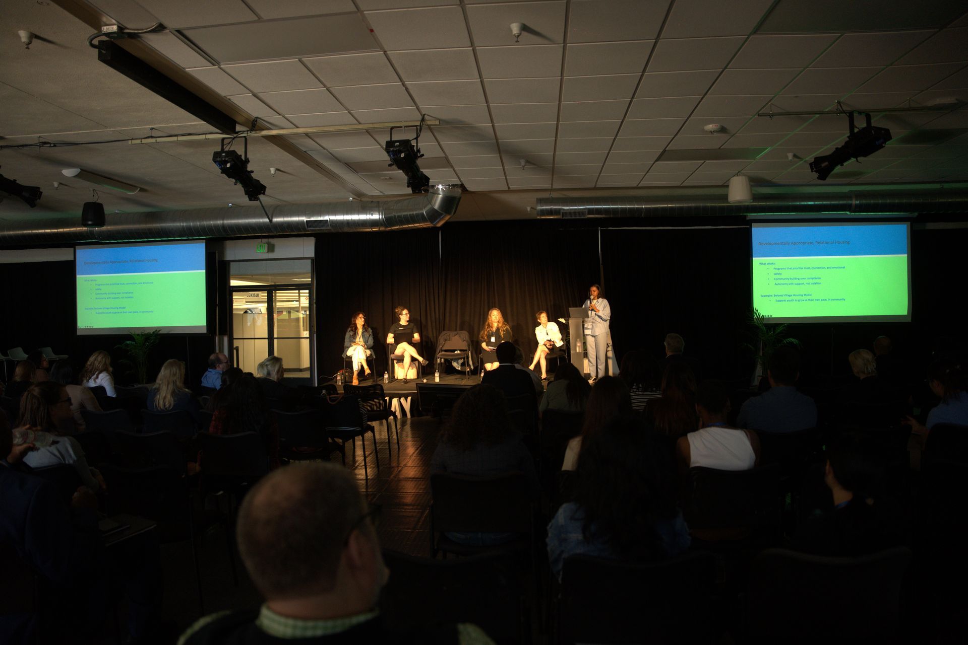 Panel discussion on stage in a dim auditorium, with two presentation screens and an audience in the foreground