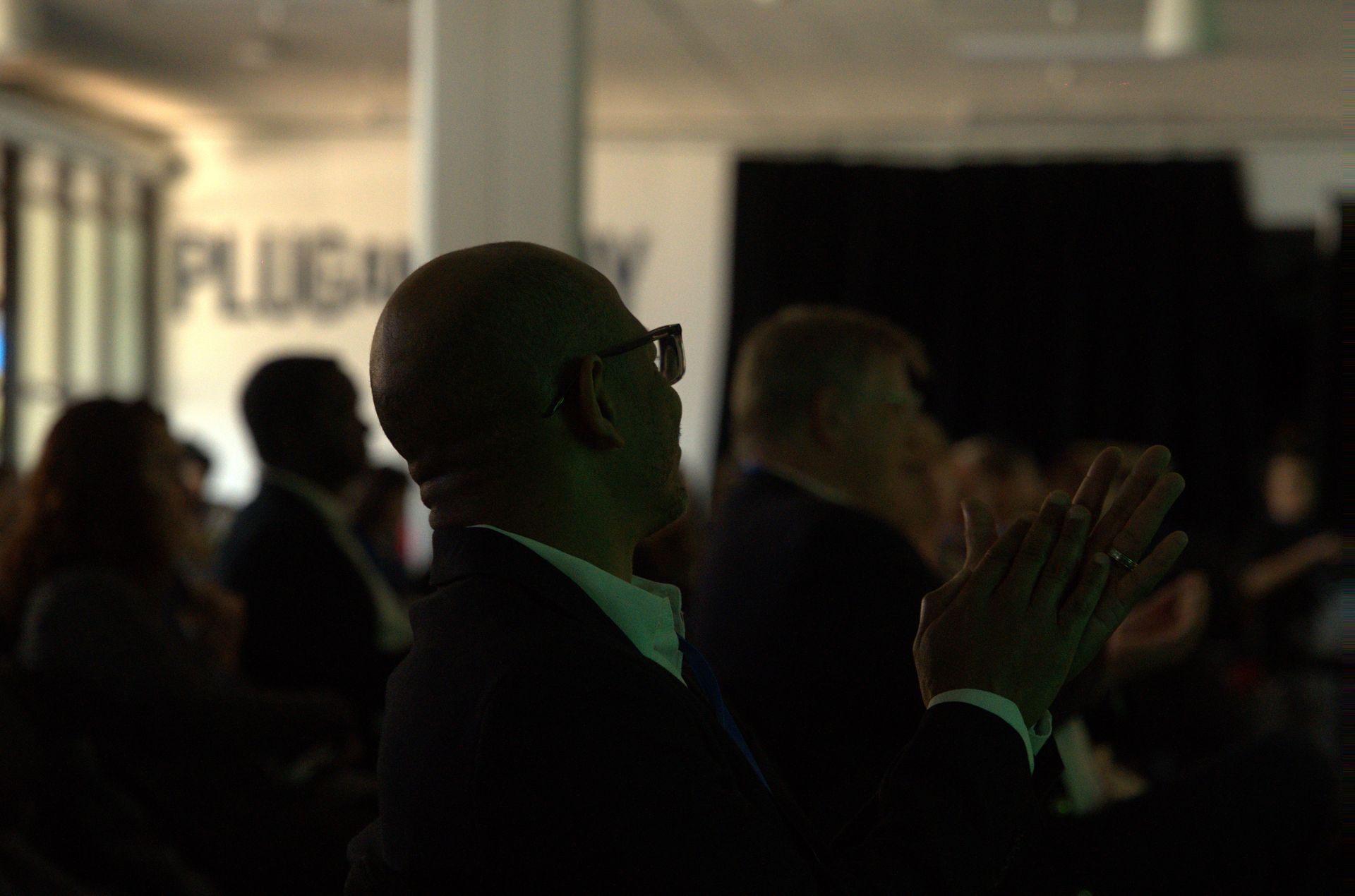 Audience applauding in a dimly lit conference room, with a green-lit man in the foreground.