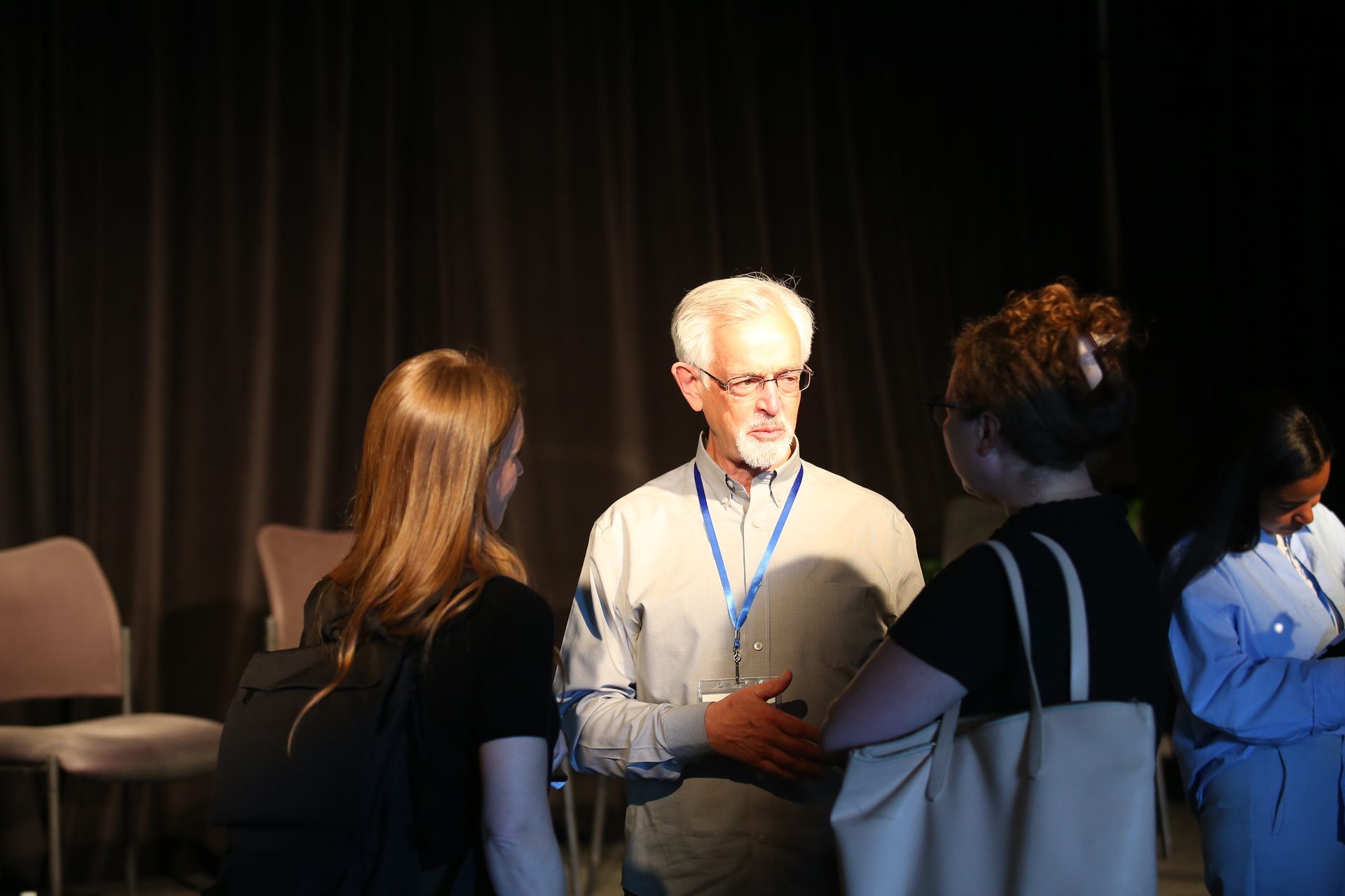Three people talking in a dimly lit event space, with one man wearing a lanyard centered in the group.