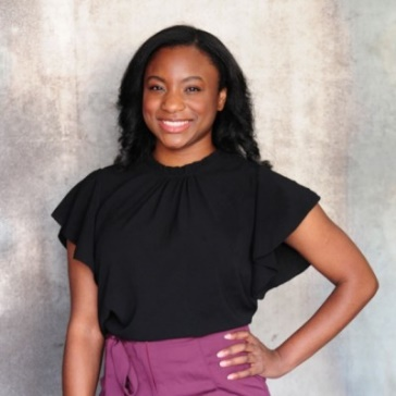 Smiling woman in a black blouse and purple skirt standing against a gray backdrop