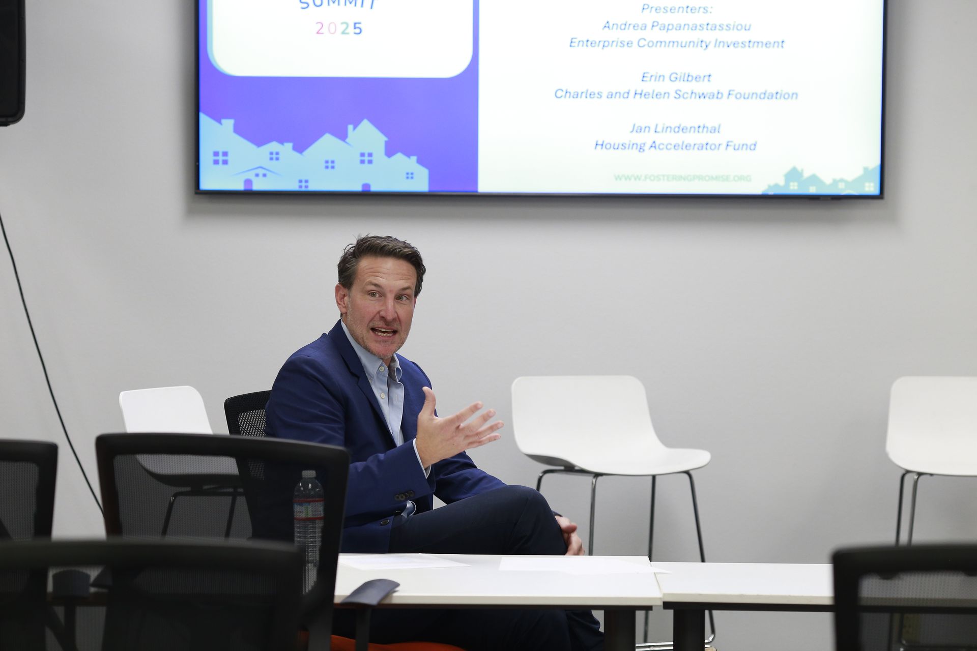 Man speaking on a stage in a conference room, seated near white chairs and a presentation screen.