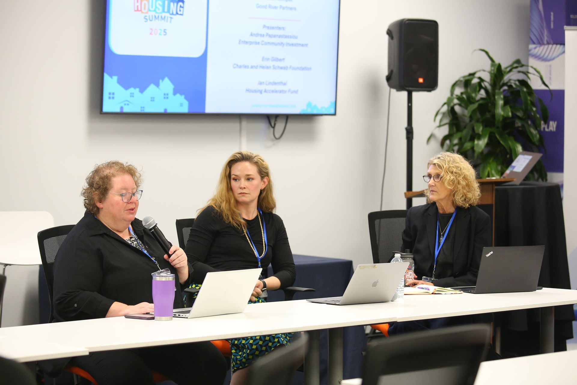 Panel discussion with three speakers seated at a table using laptops and microphones in a meeting room.