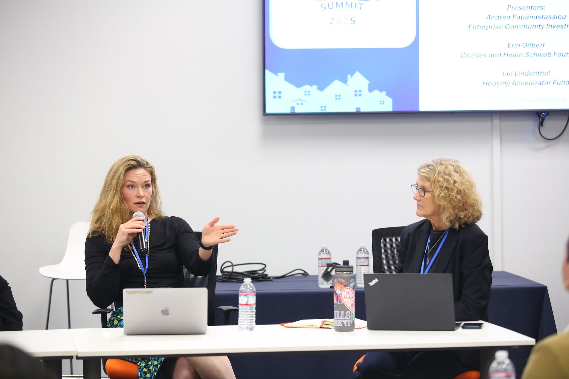 Two speakers at a panel table, one gesturing while speaking into a microphone, with laptops and a presentation screen behind them.