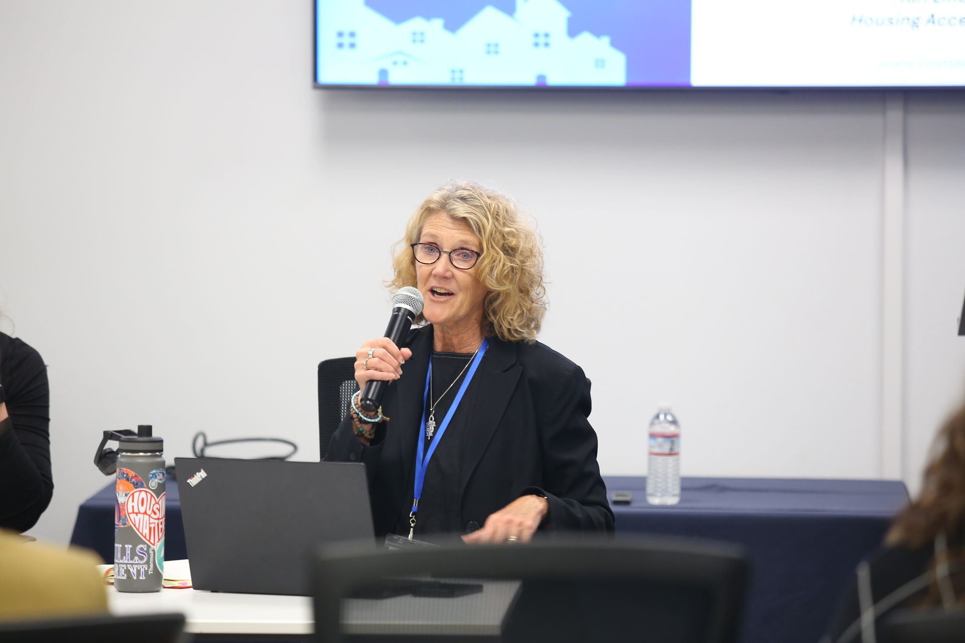 Speaker at a podium holding a microphone in a conference room, with a laptop and water bottle nearby.