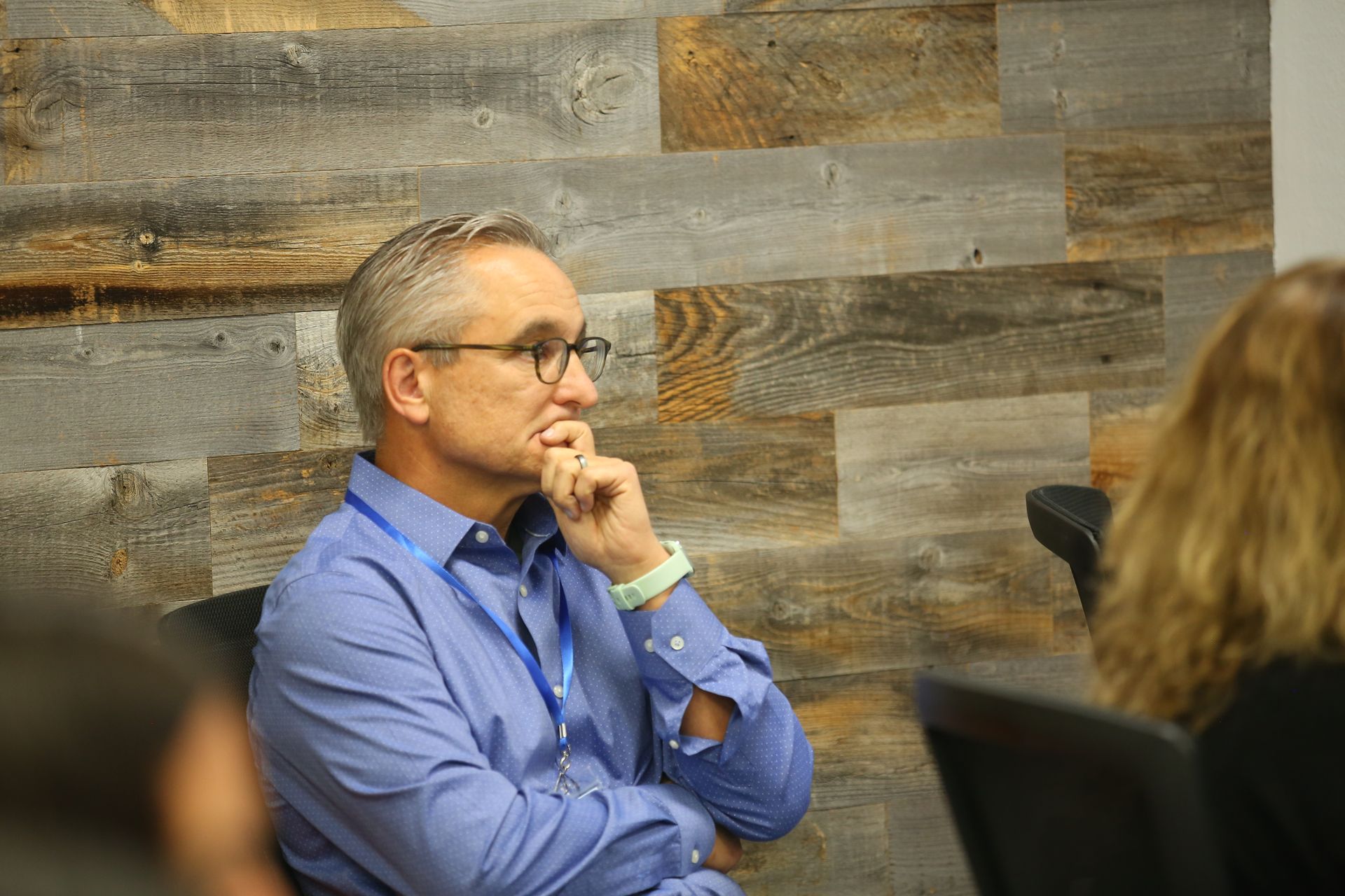 Man in blue shirt and glasses listening thoughtfully at a meeting, seated against a wood-paneled wall