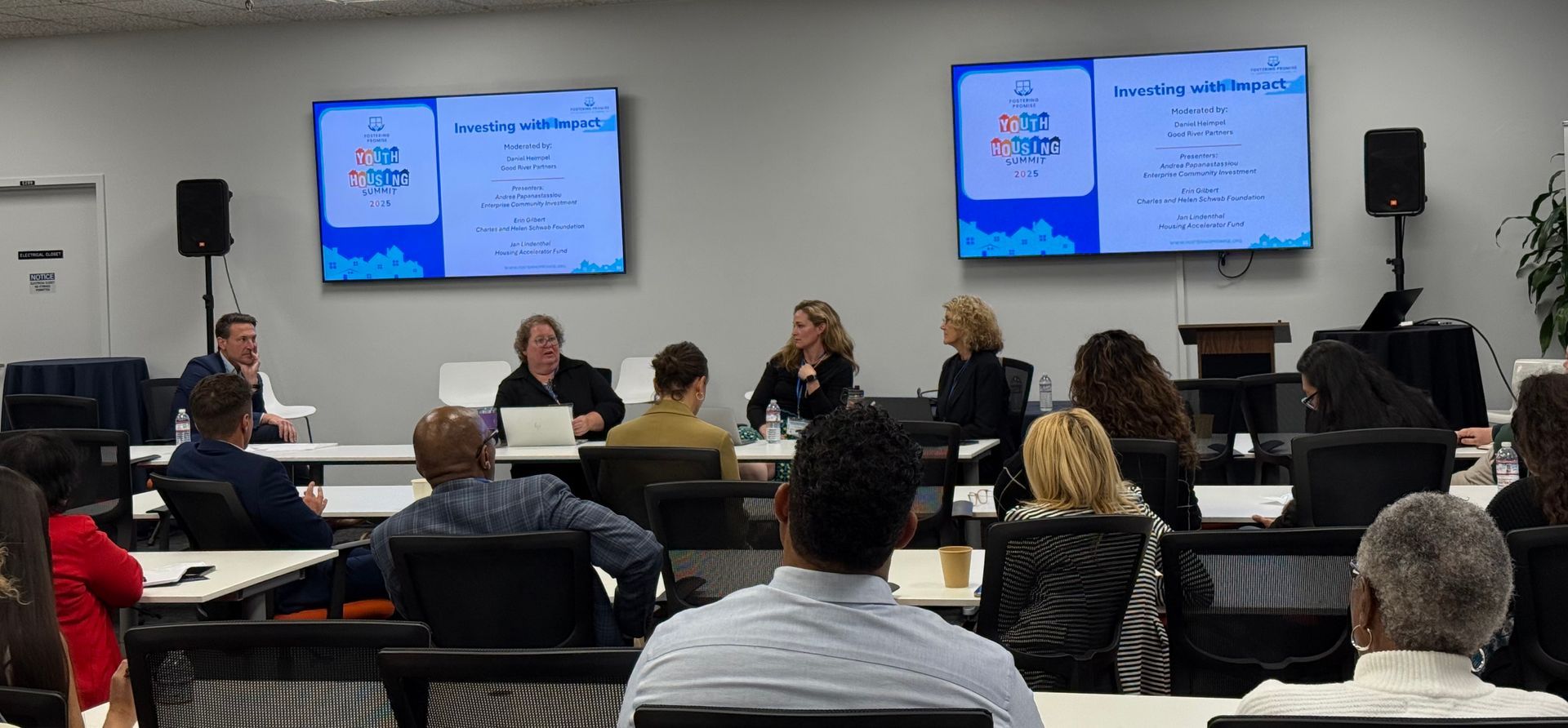 Audience seated in a conference room watching a panel discussion on two wall-mounted screens