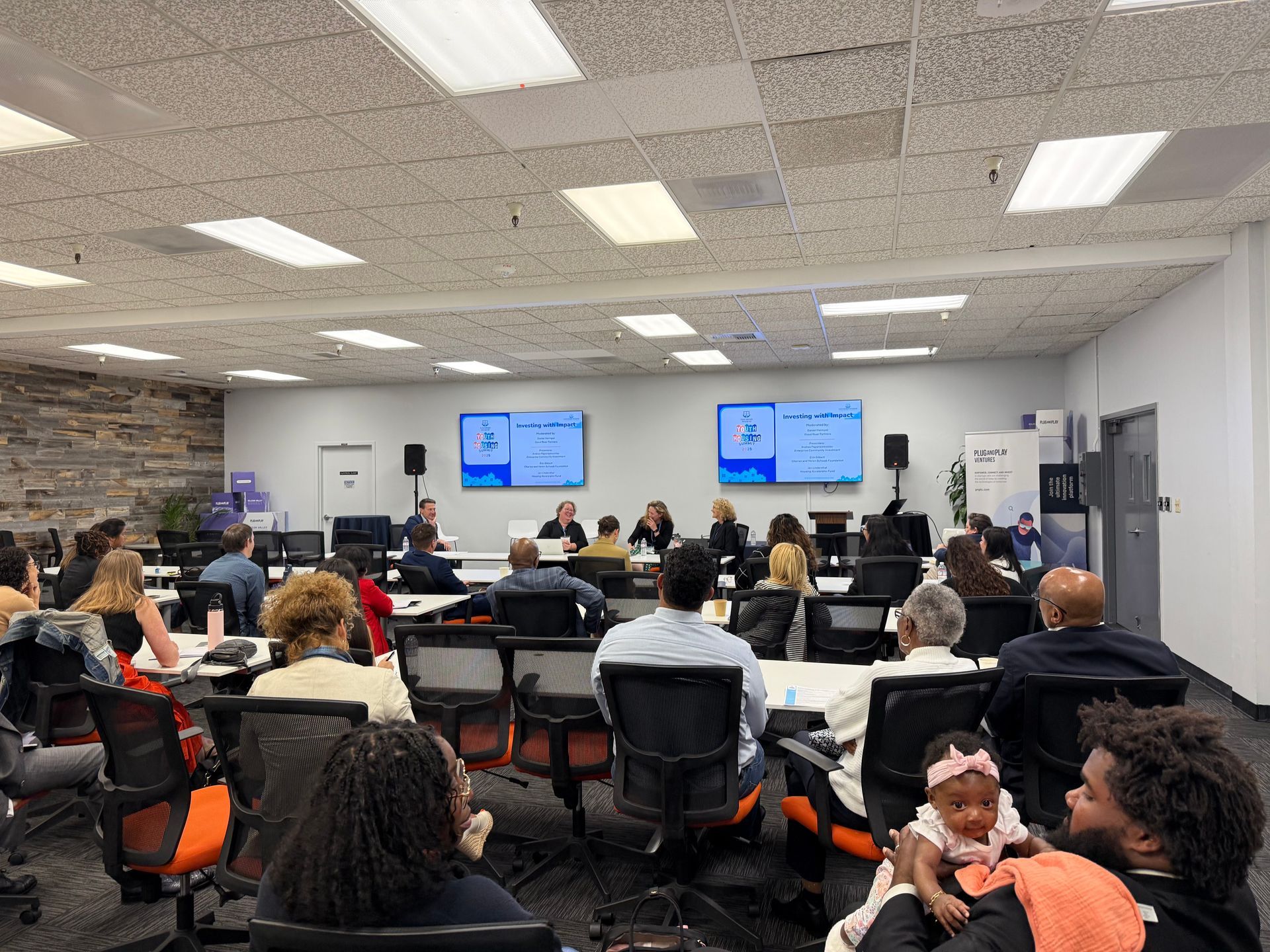 Conference room classroom with people seated at desks facing two presentation screens at the front.