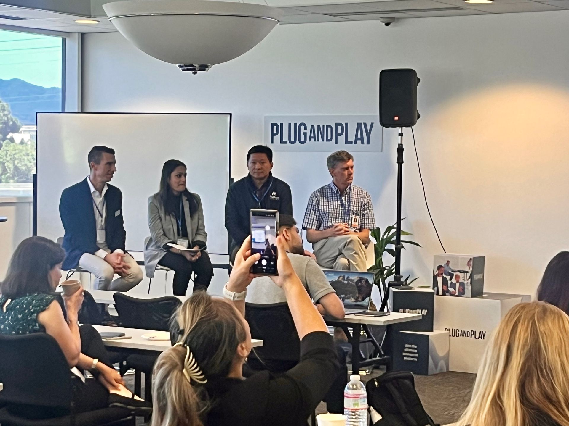 Panel discussion at Plug and Play with four speakers on stage and an audience member filming.