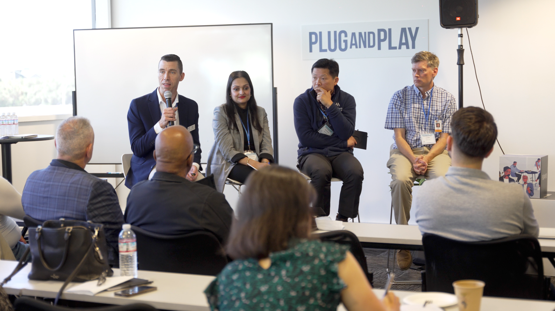 Panel discussion in a conference room with four speakers seated on stage and attendees watching.