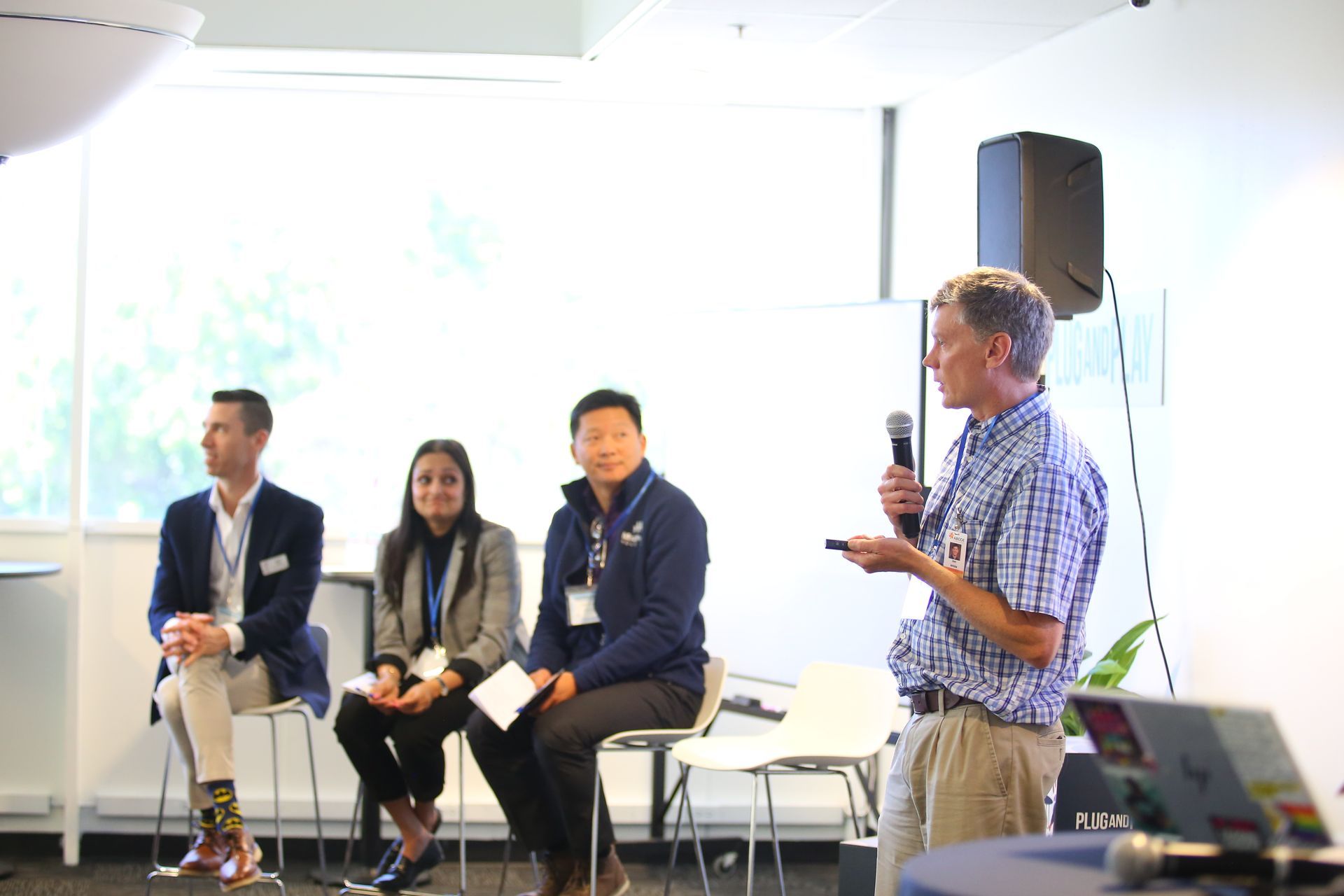 Panel discussion in a bright room, with three seated speakers and one standing man holding a microphone.