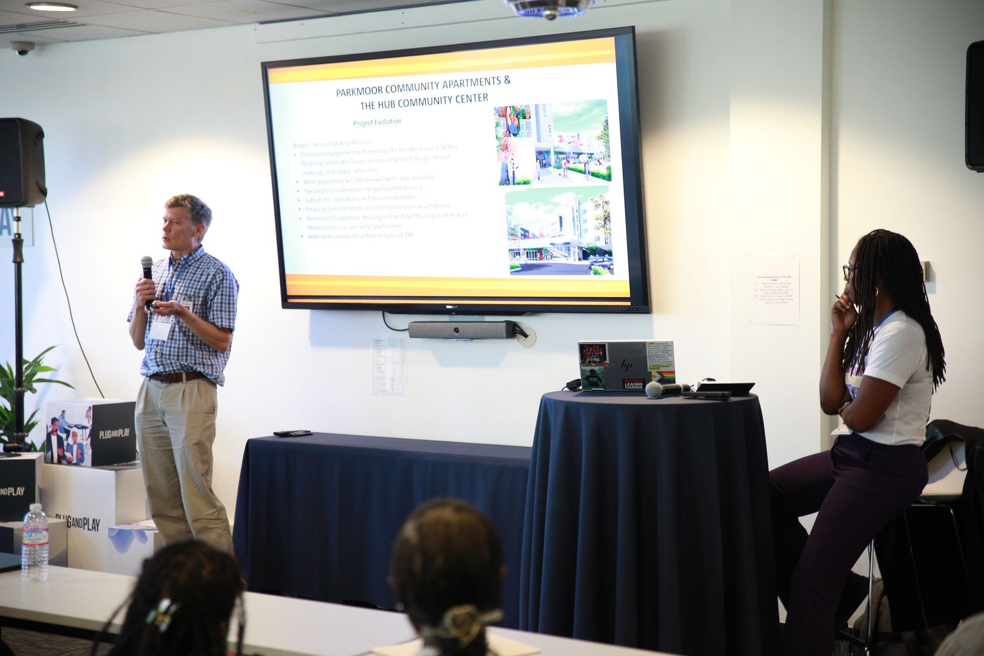 Presenter speaking beside projected slide in a conference room, with audience seated nearby.
