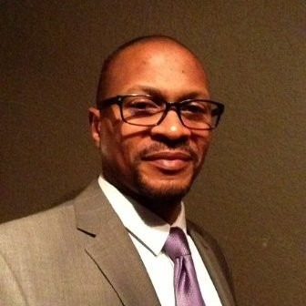 Portrait of a man in a gray suit and purple tie, wearing glasses and smiling against a dark background
