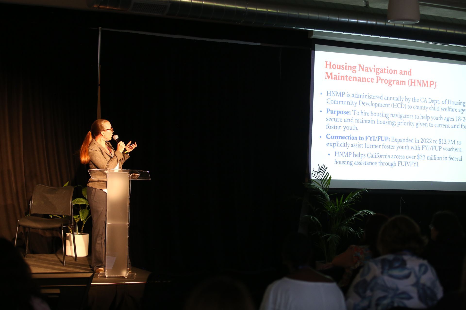 Speaker at podium presenting slides on a projector screen in a dark conference room.