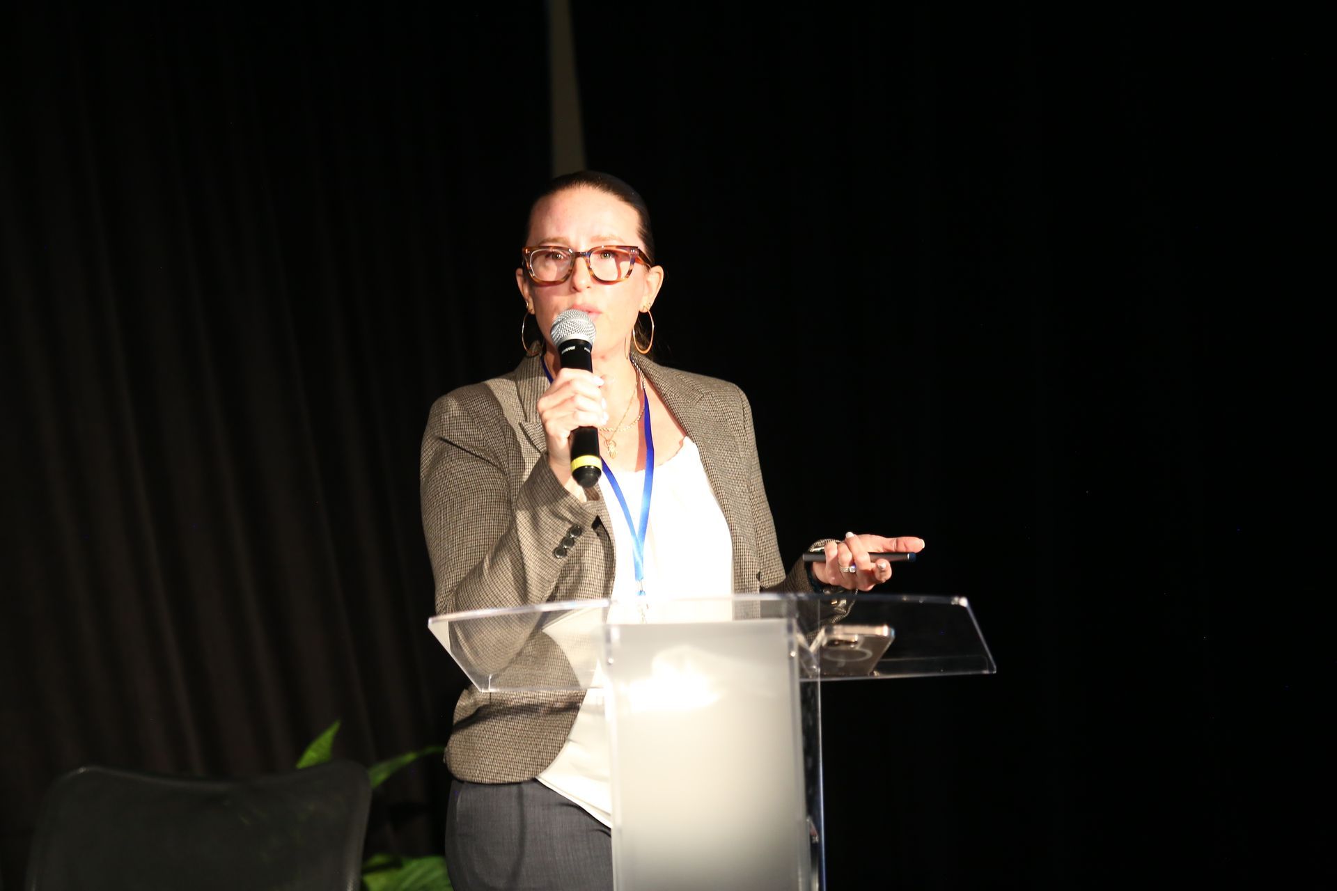 Person speaking at a podium on stage with a microphone, wearing glasses and a blazer.