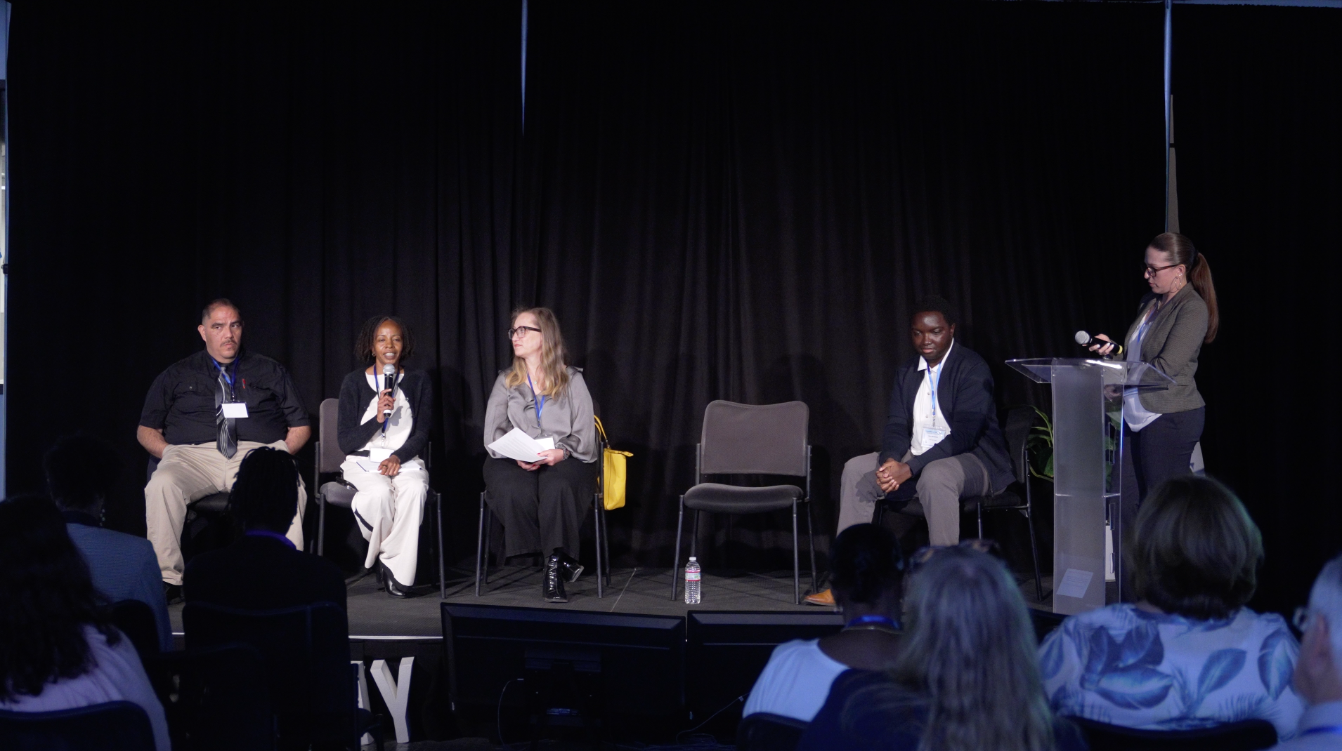 Panel discussion on a stage with four seated speakers, a standing moderator, and audience silhouettes in front