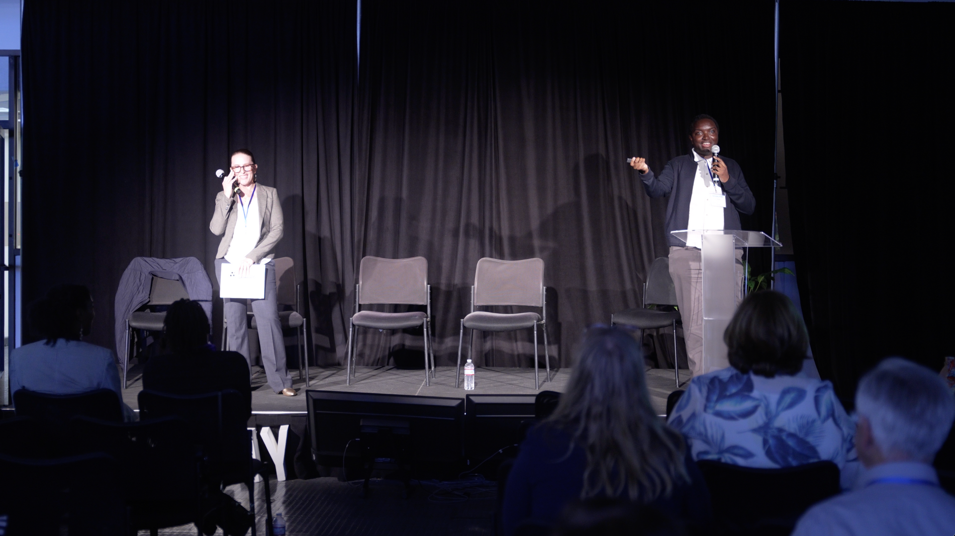 Two speakers on a stage with black curtains, one seated audience visible in foreground