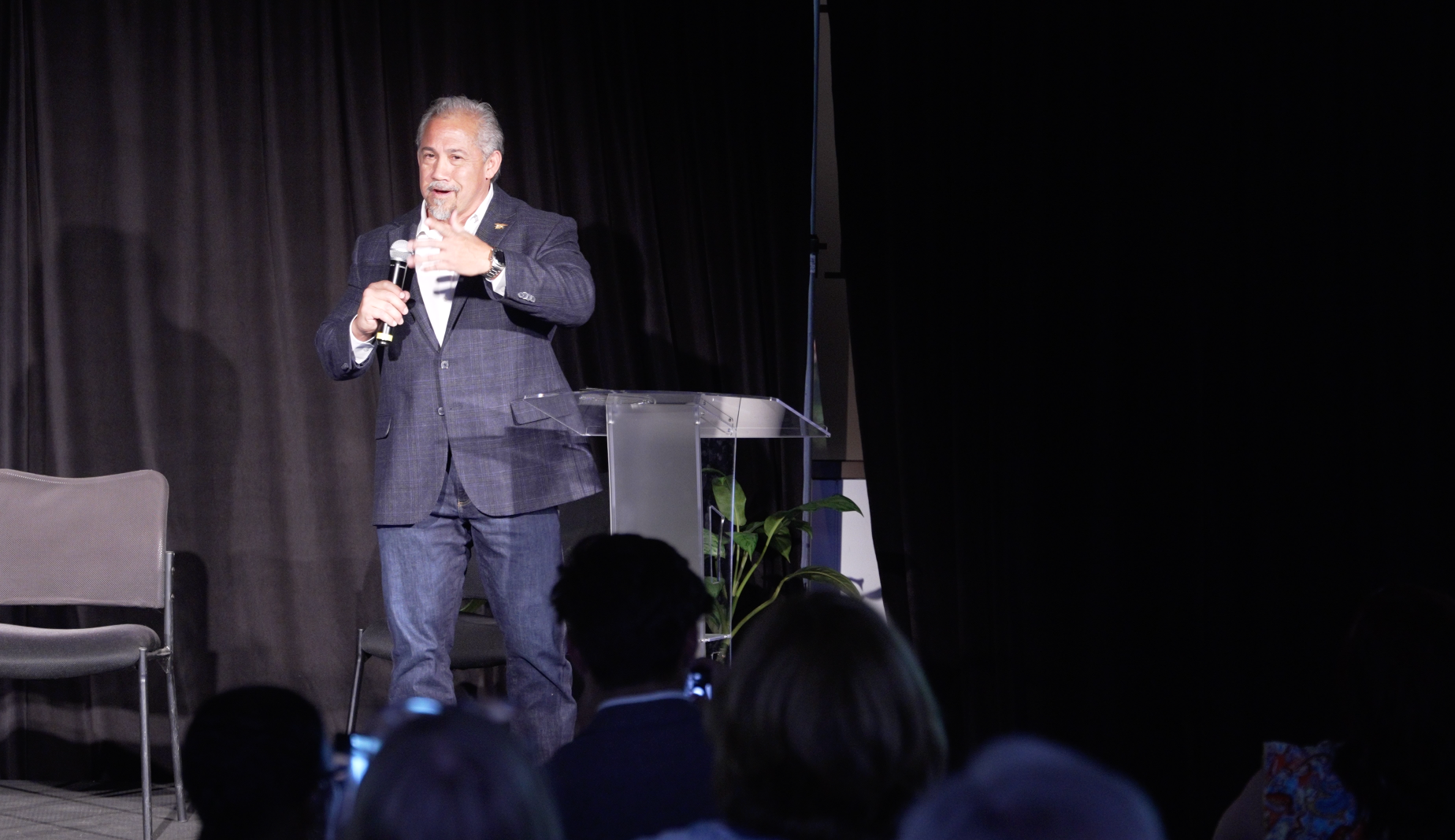 Speaker on stage holding a microphone, with a table, chair, and audience silhouettes in front.