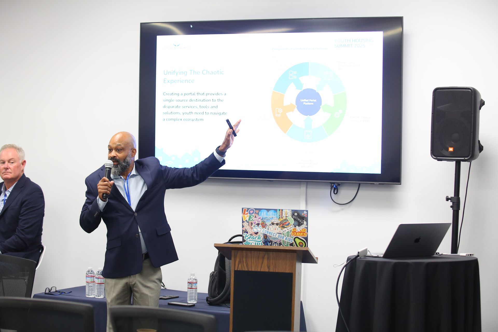 Presenter with microphone gestures beside a wall-mounted screen during a conference talk