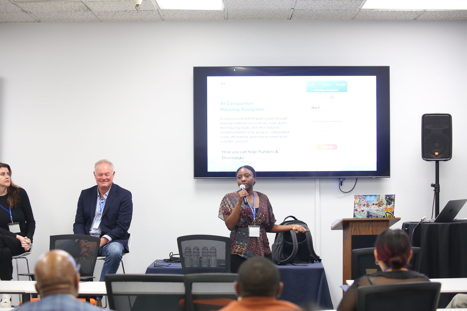 Panel discussion in a conference room with speakers seated before a presentation screen and audience.