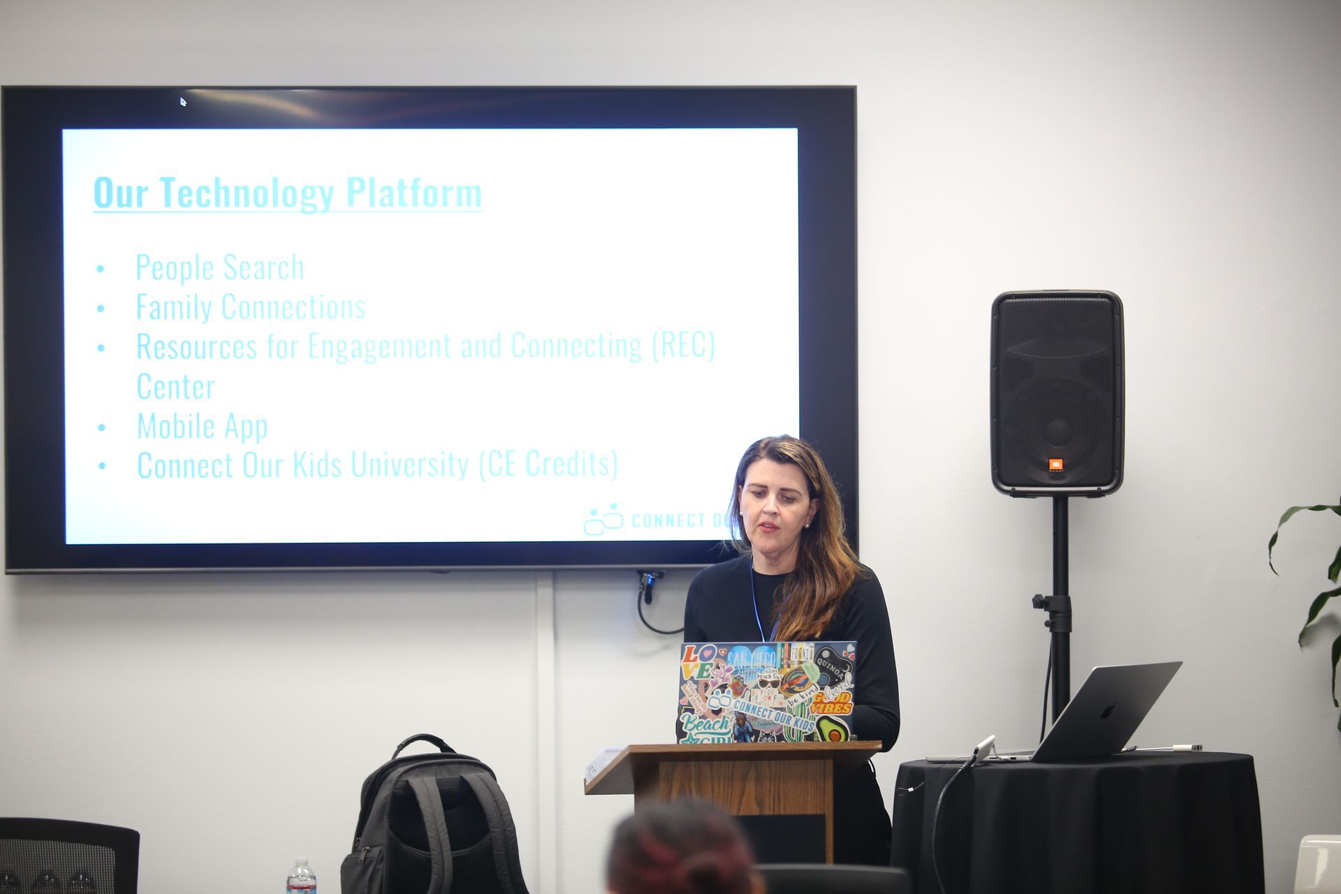 Woman presenting at a podium with a laptop in front of a screen, beside a speaker in a conference room