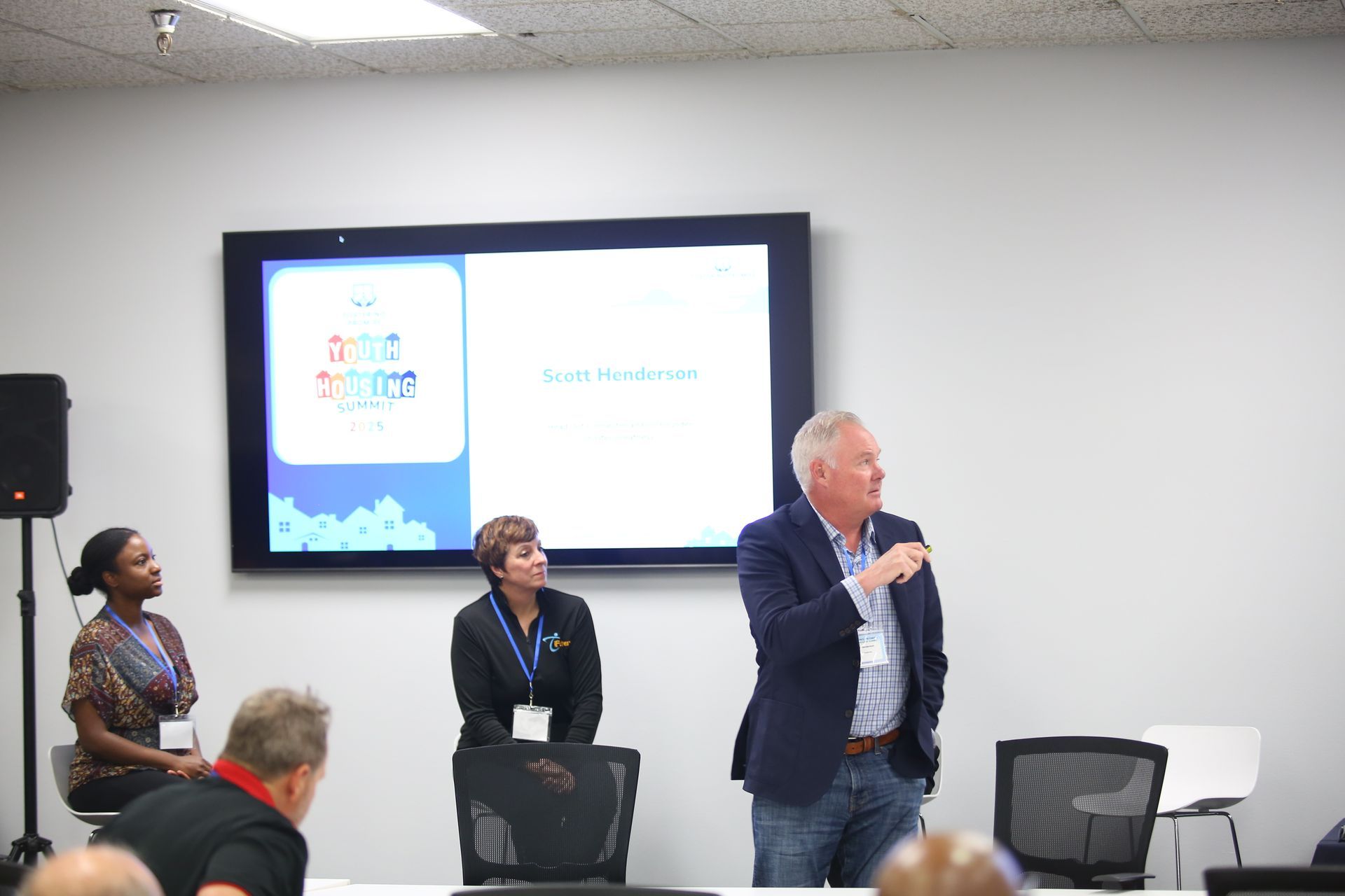 Presenter speaking in a meeting room beside a wall-mounted screen, with attendees seated nearby