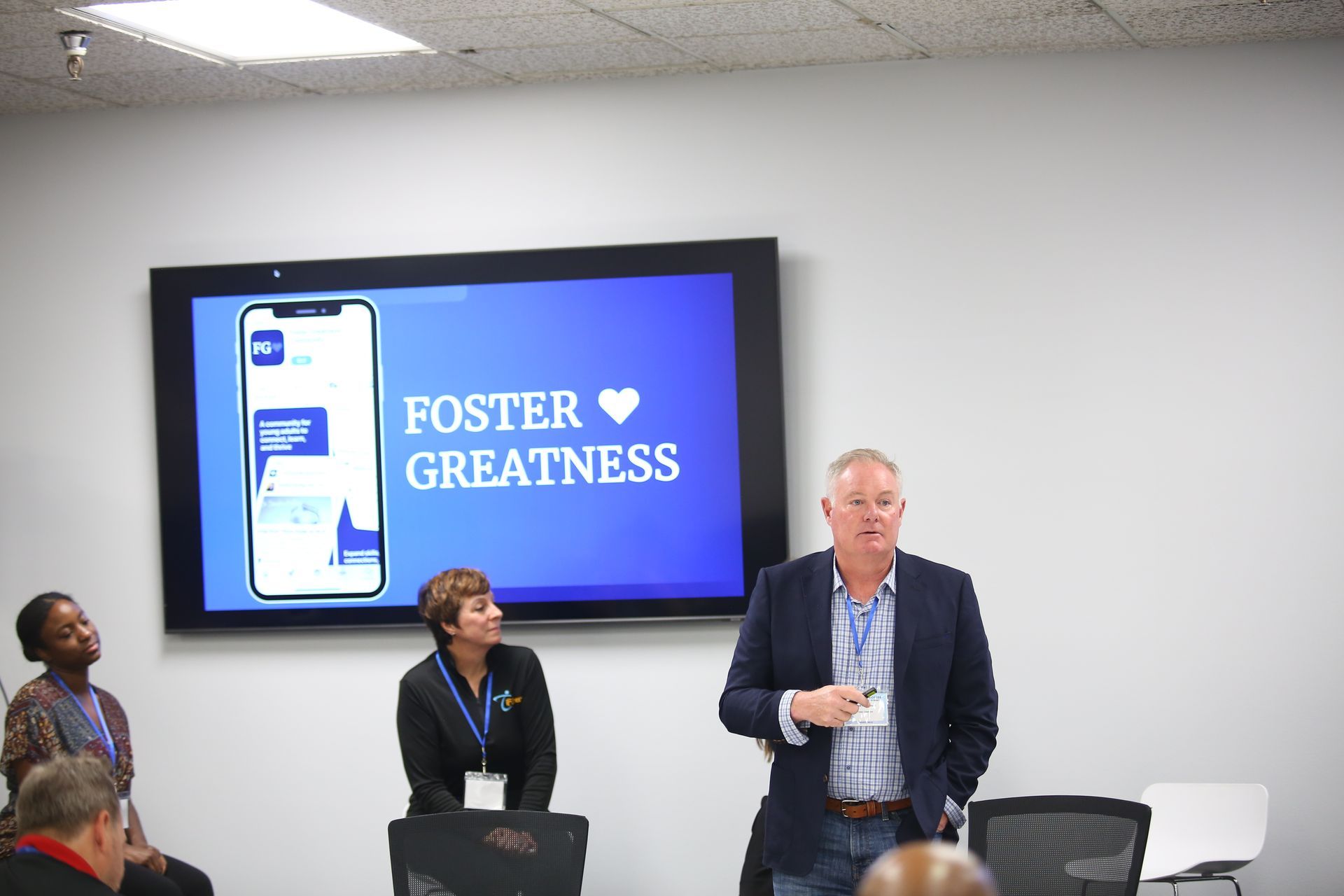 Man speaking in a conference room beside a screen reading “Foster Greatness” with a phone graphic.