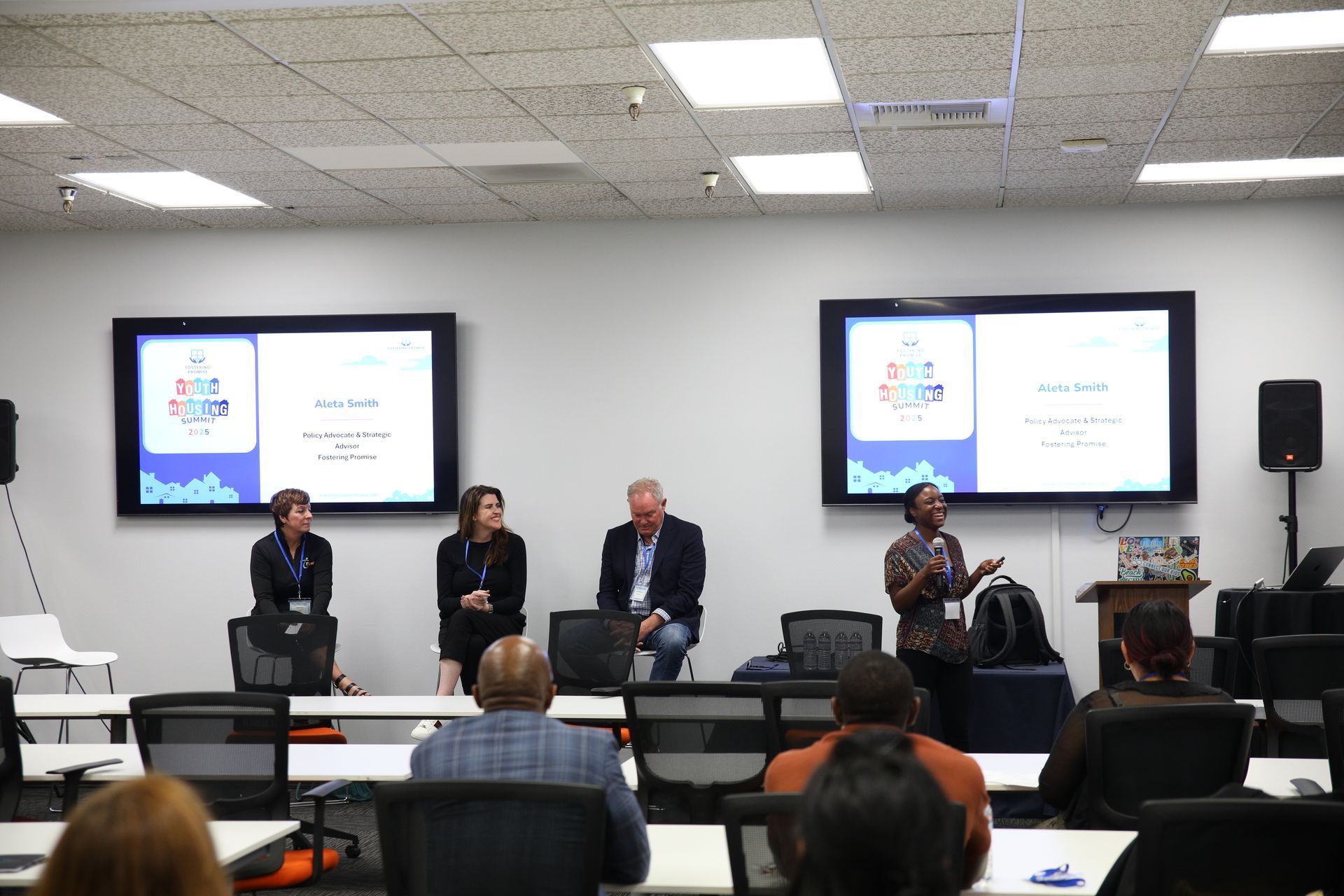 Panel discussion in a lecture room, with four speakers seated before two presentation screens.