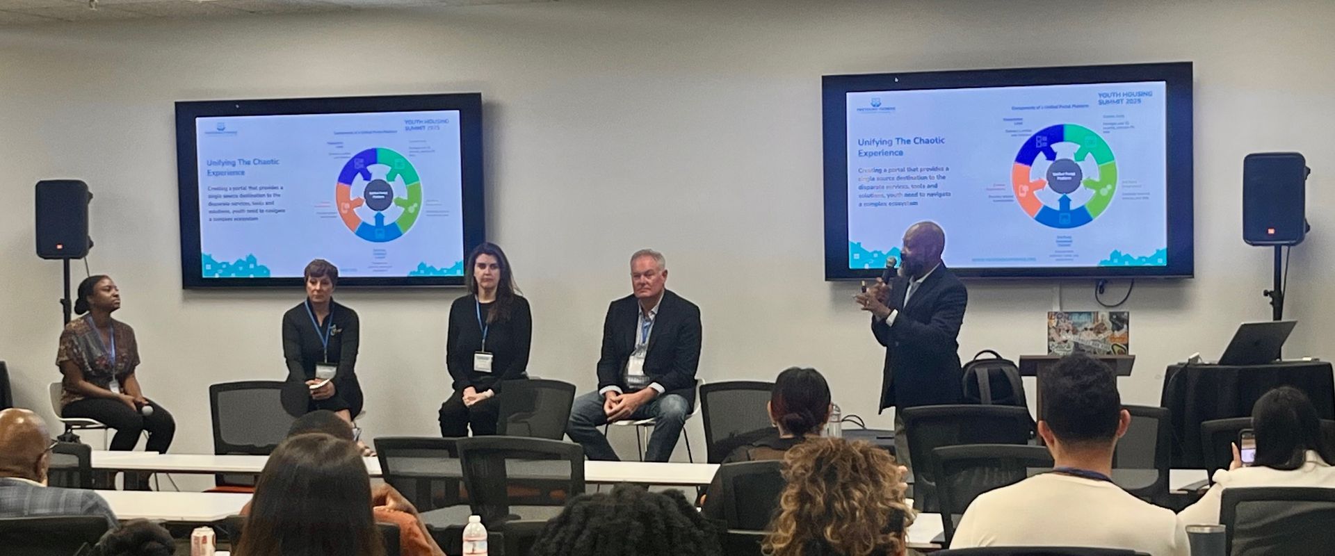 Panel discussion on stage with five speakers, two presentation screens, and an audience in the foreground