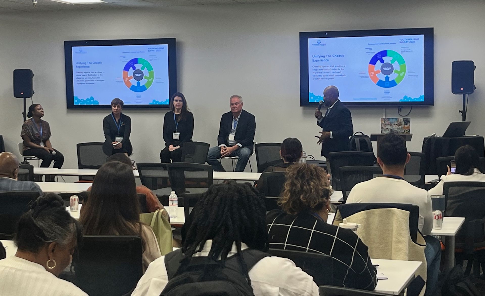 Panel discussion in a classroom, with speakers on stage and audience seated facing two presentation screens.