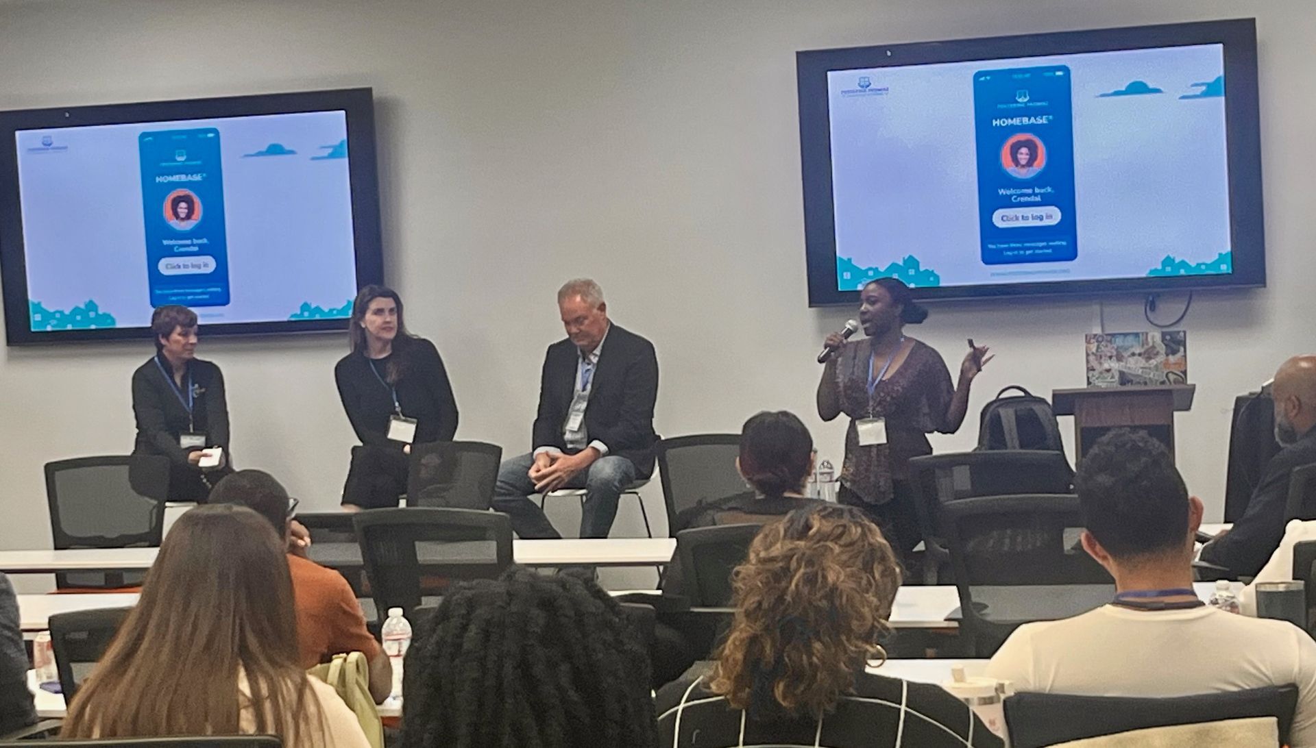 Panel discussion in a classroom, with four speakers seated before two screens showing a presentation slide.