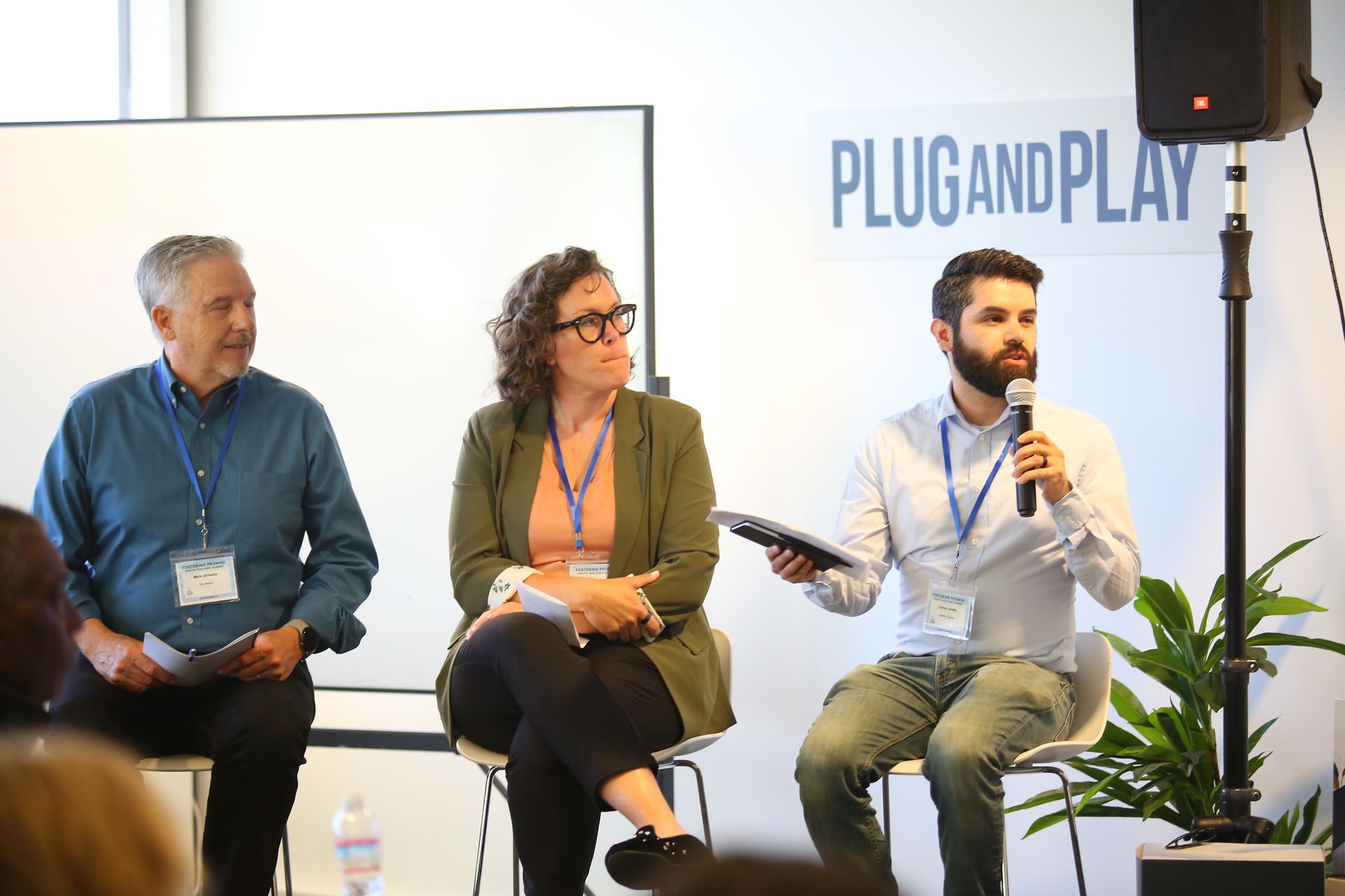 Three speakers seated on a stage during a panel discussion, with a “PLUG AND PLAY” backdrop.