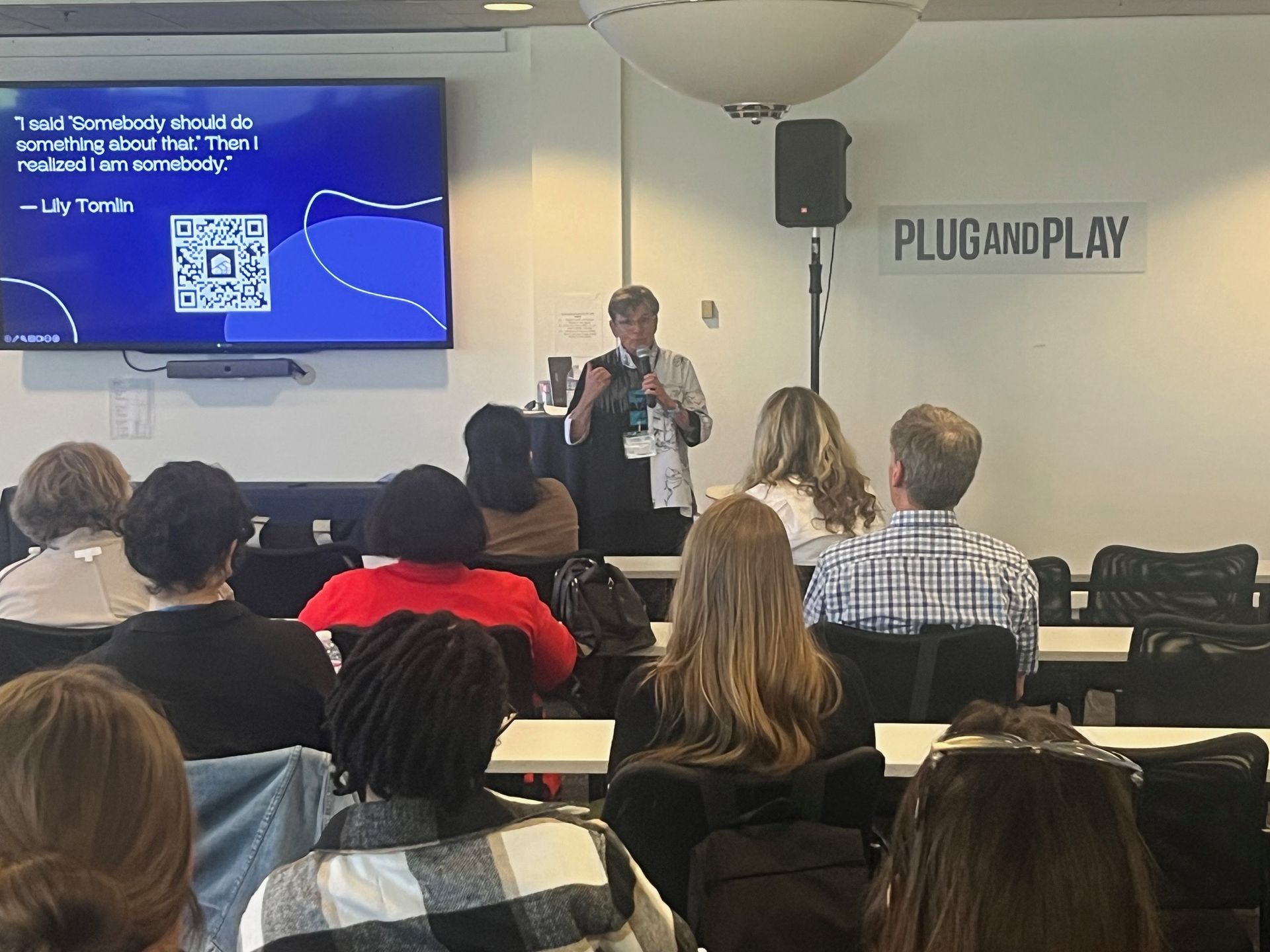 Speaker presenting at a tech event to an audience in a room with a “PLUG AND PLAY” sign and slide screen.