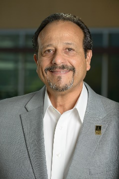 Smiling man in a light gray blazer and white shirt against a blurred indoor background