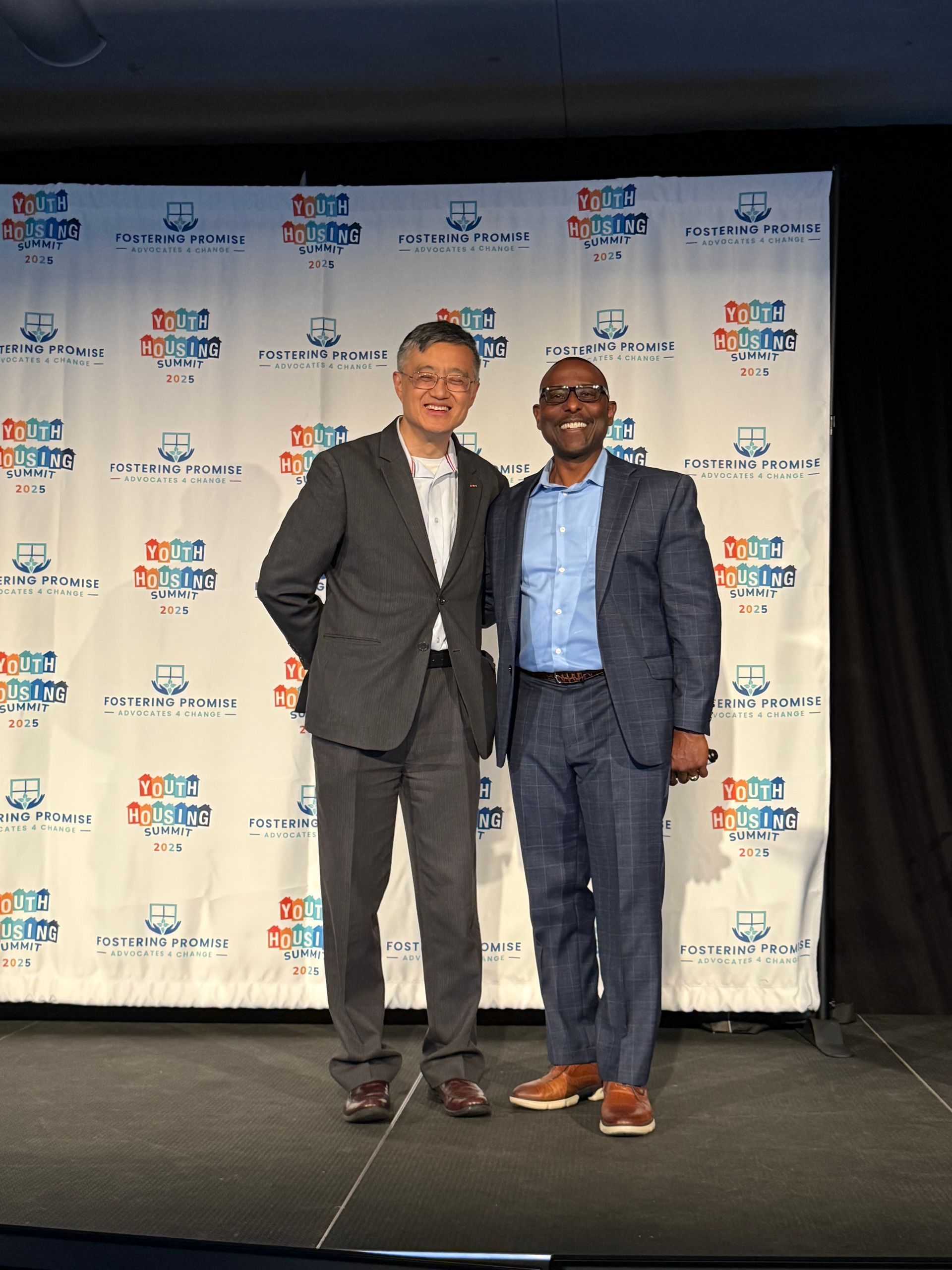 Two men in suits posing on a stage in front of a logo backdrop.