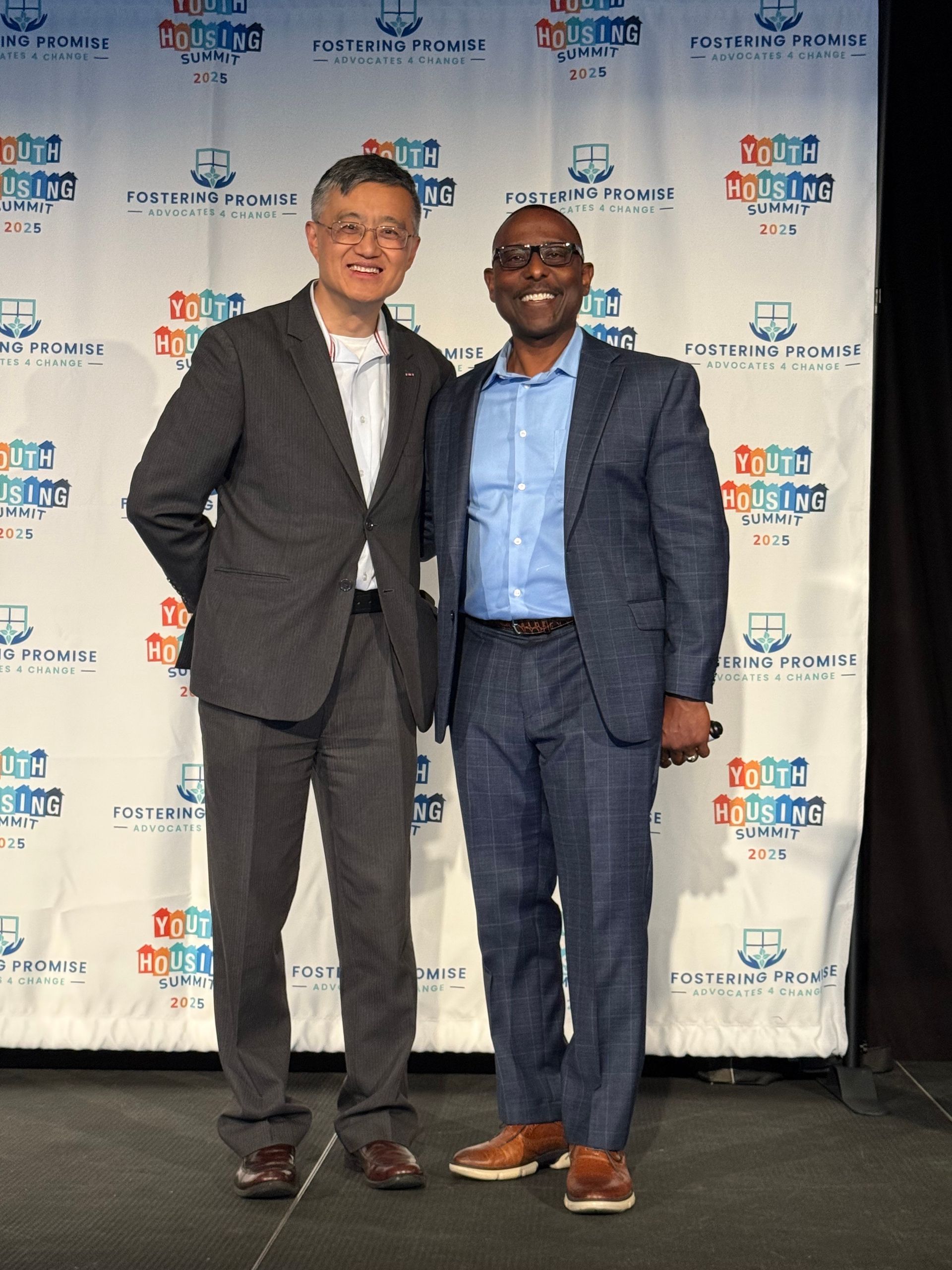 Two men in suits posing together at an event with a branded step-and-repeat backdrop