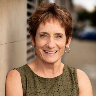 Smiling woman in an olive green sleeveless top standing outdoors by a wall.