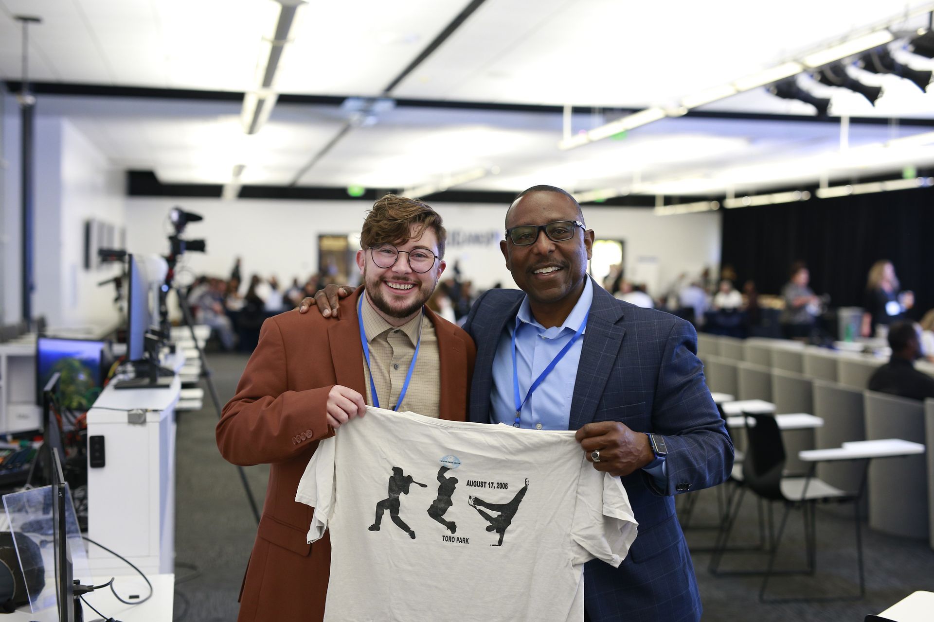 Two men in an office hold up a beige T-shirt with a black graphic, smiling at the camera