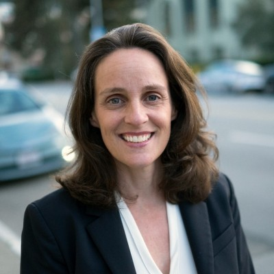 Woman in a dark blazer smiling outdoors on a city street, with blurred cars in the background