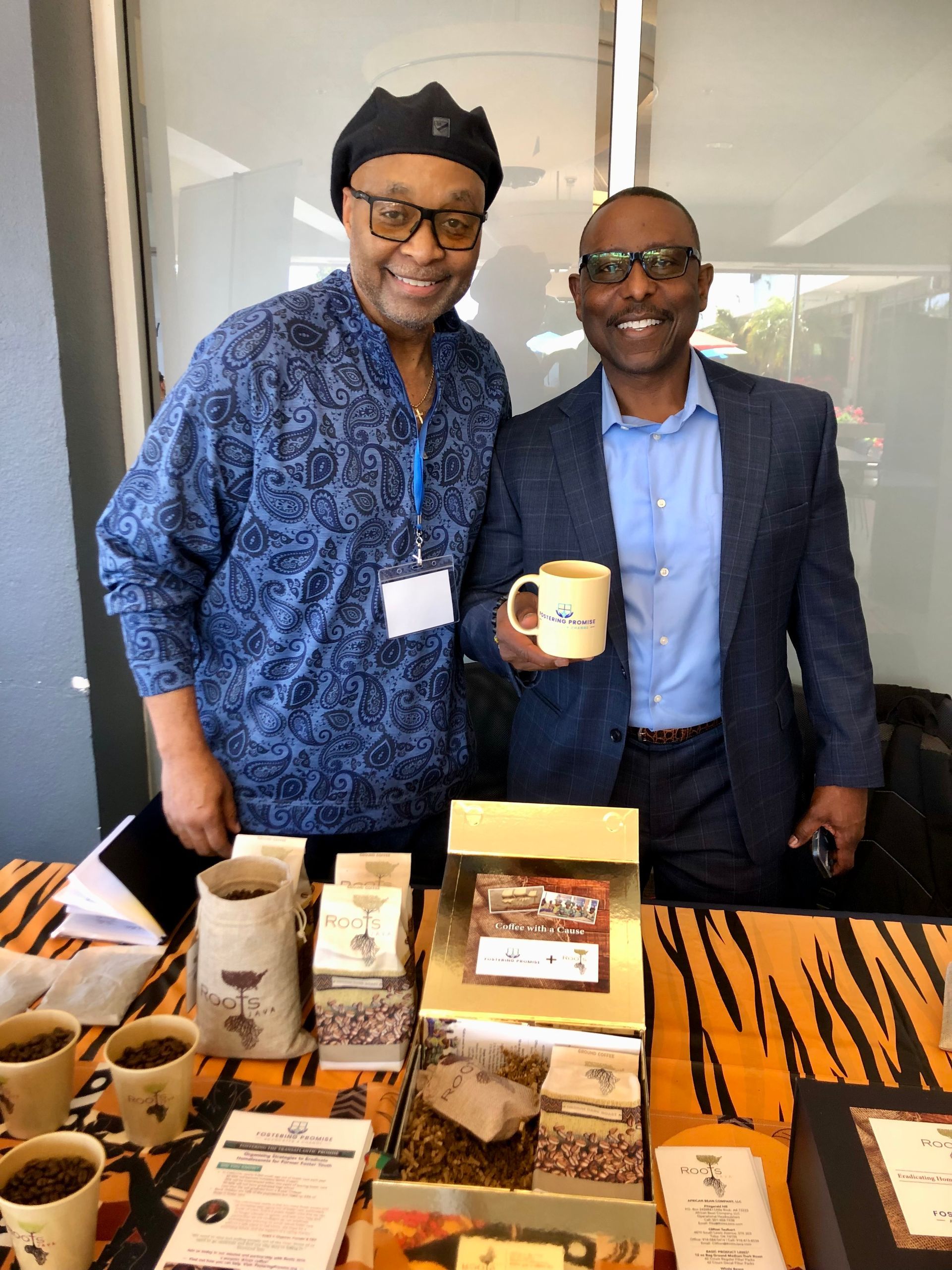 Two men smiling at a table display of packaged goods, one holding a mug.
