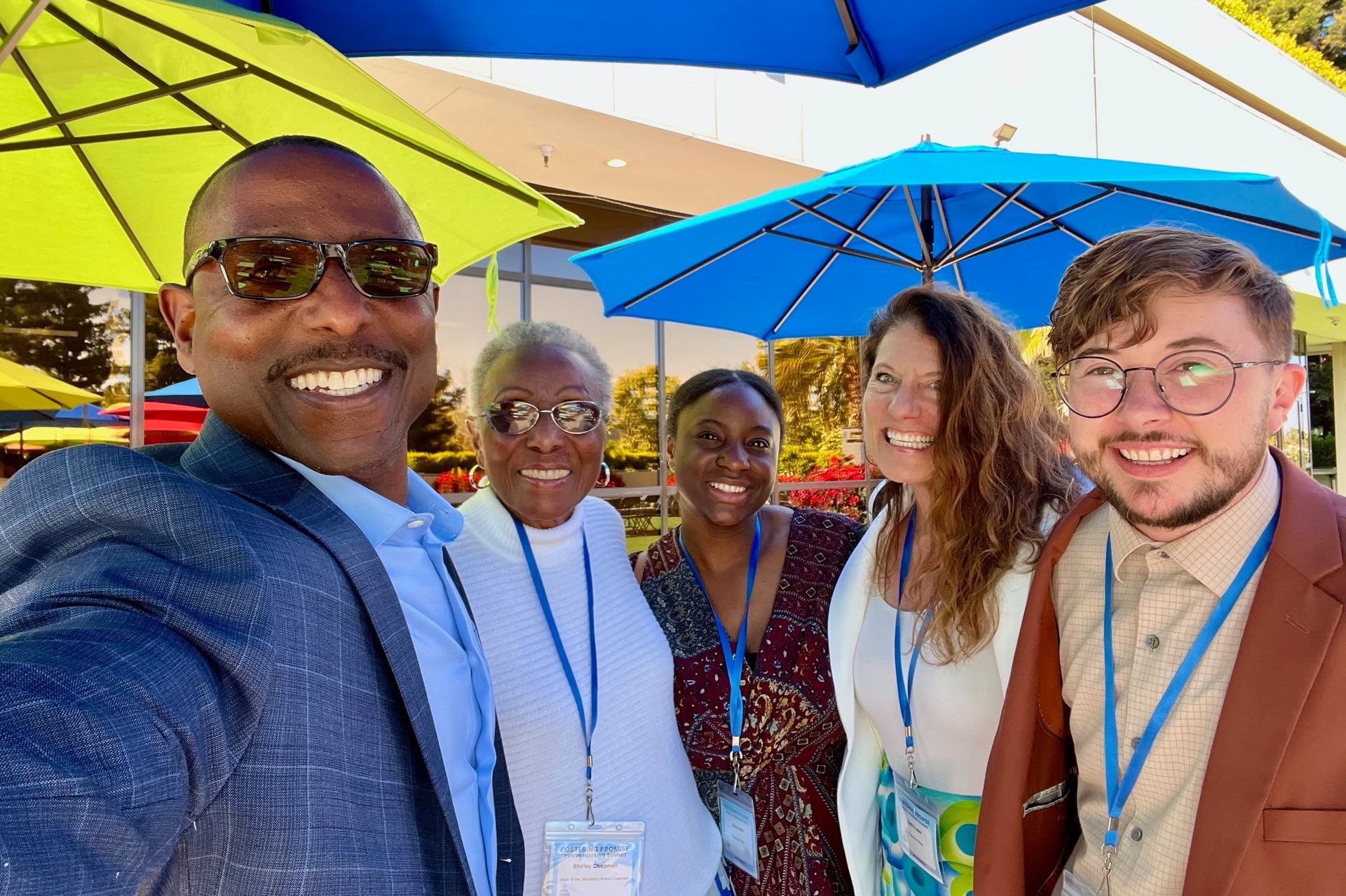 Five smiling people at an outdoor event under bright blue and yellow umbrellas
