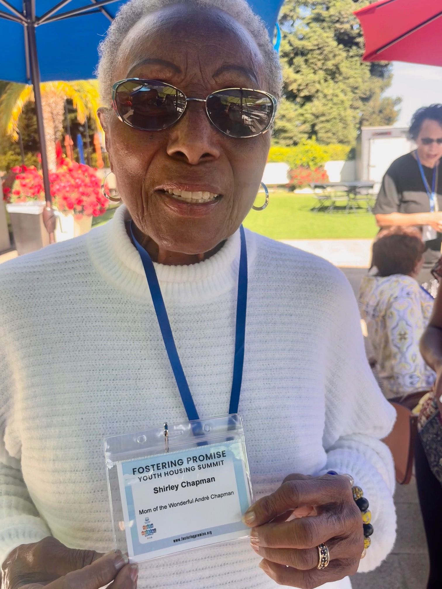 Person in sunglasses holds up a name badge outdoors at an event.