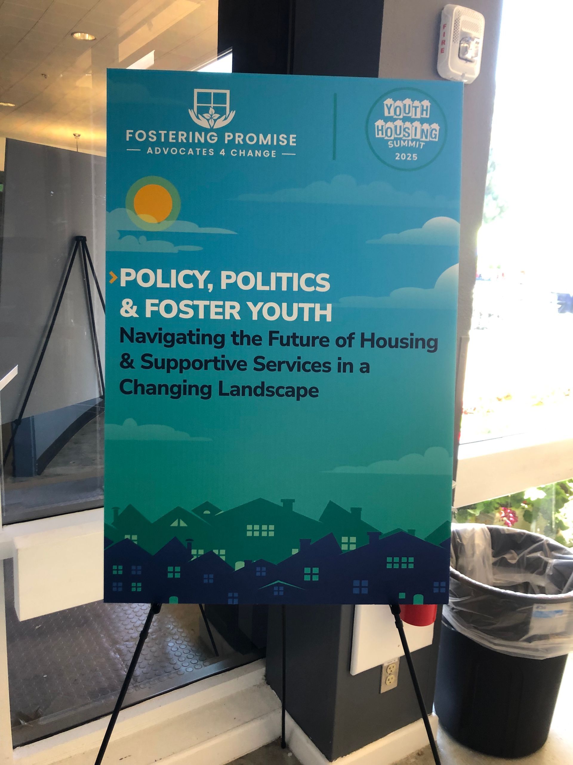 Teal event sign on an easel about policy, politics and fostering youth, in a room by a window